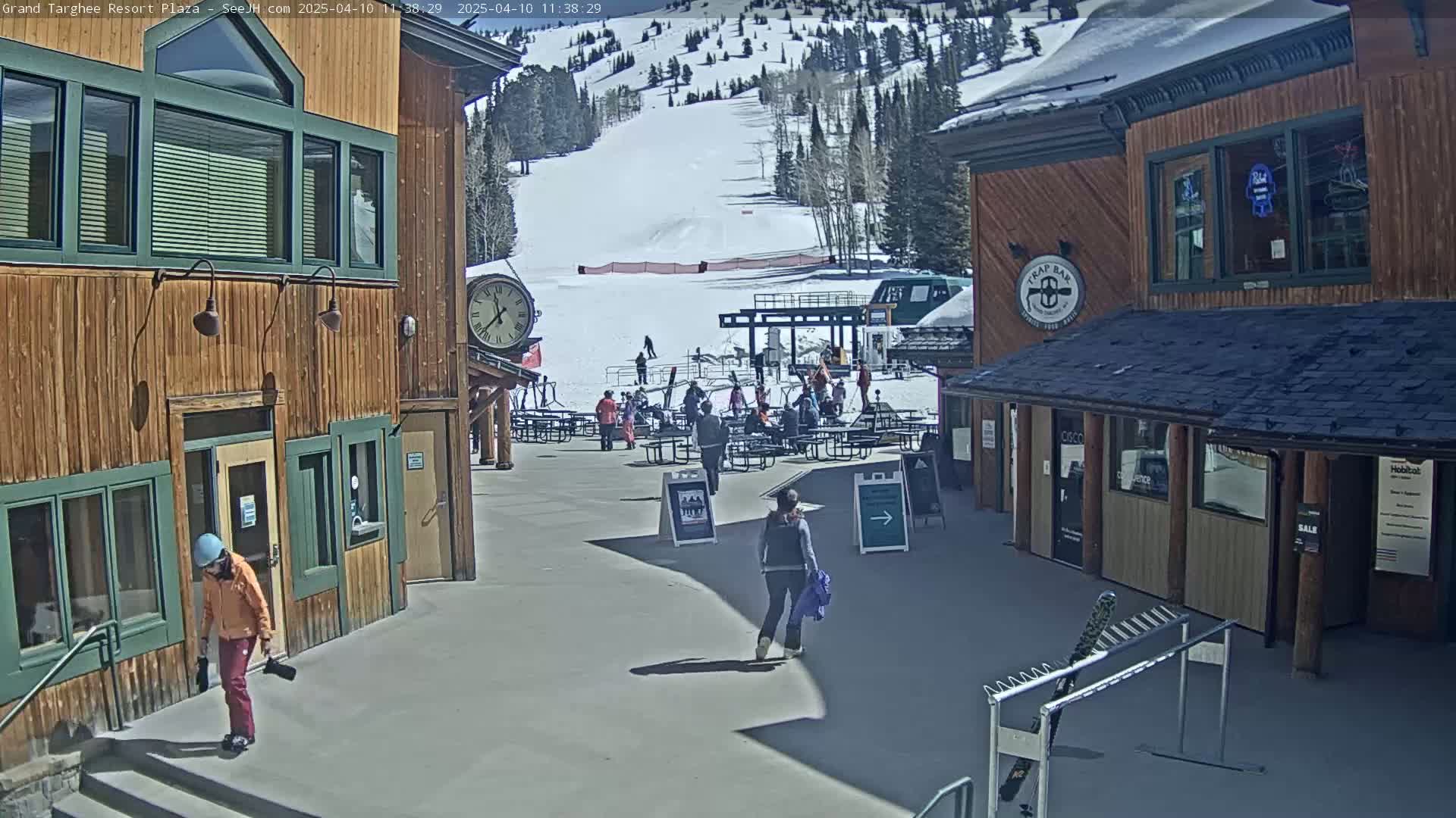 A sunny plaza area with wooden buildings, a few people walking around, and a ski slope visible in the background.