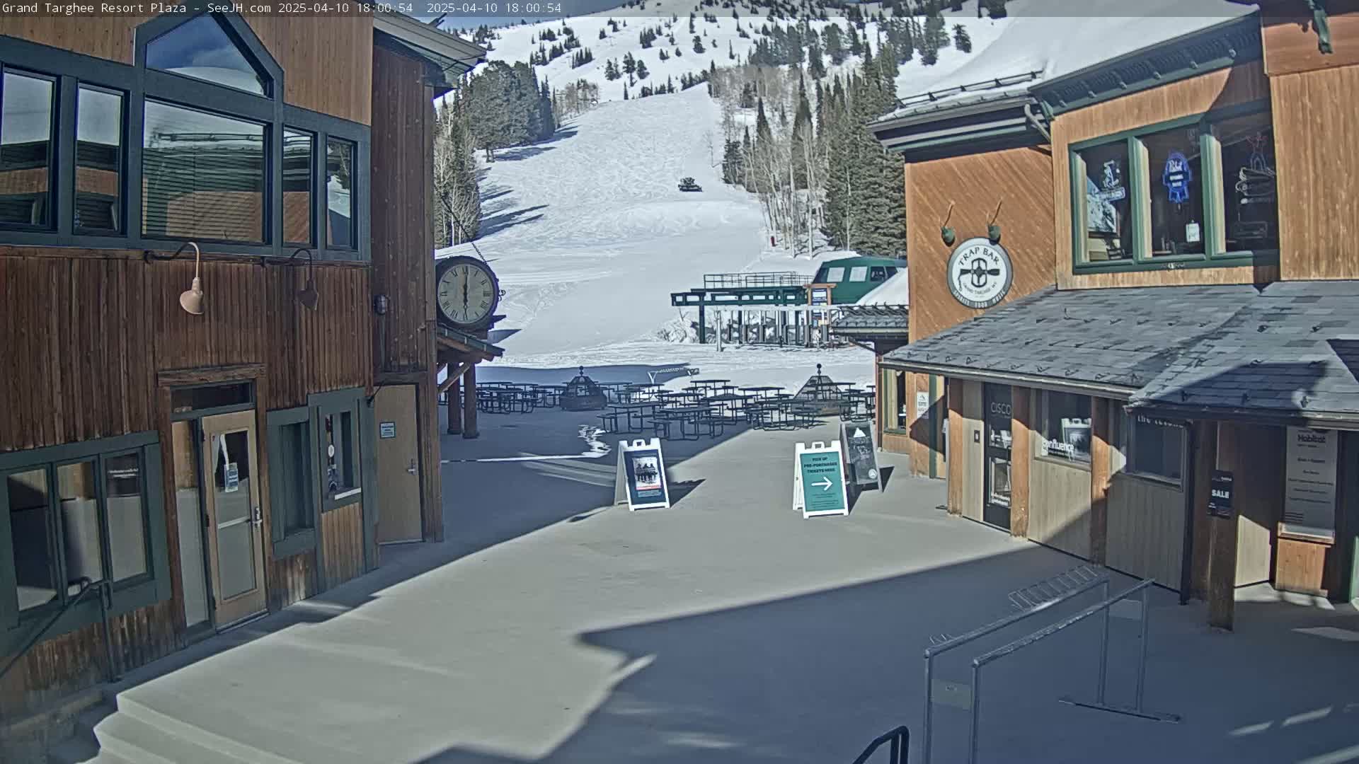A plaza with wooden buildings, snow-covered ground, and picnic tables sits in front of a ski slope on a sunny day.