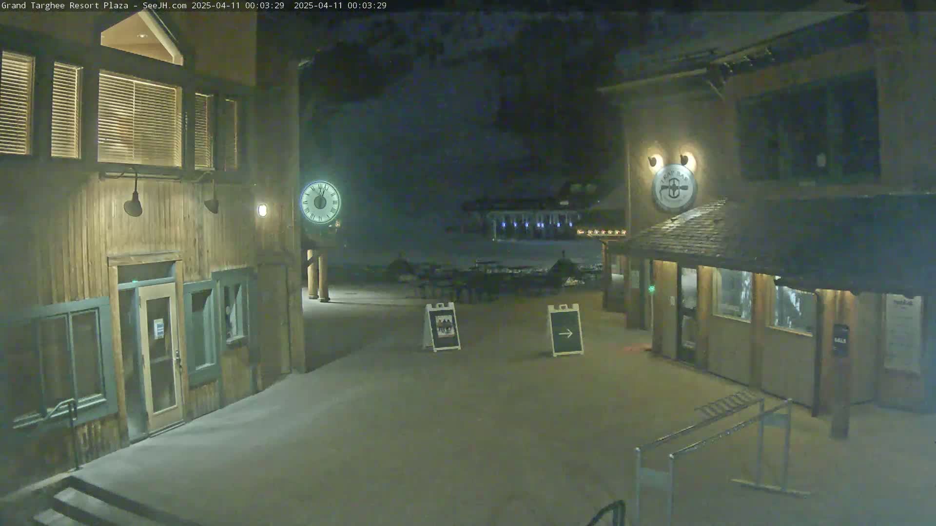 A snow-covered plaza at night, illuminated by lights, features two wooden buildings with signage, a clock, and a few directional signs.