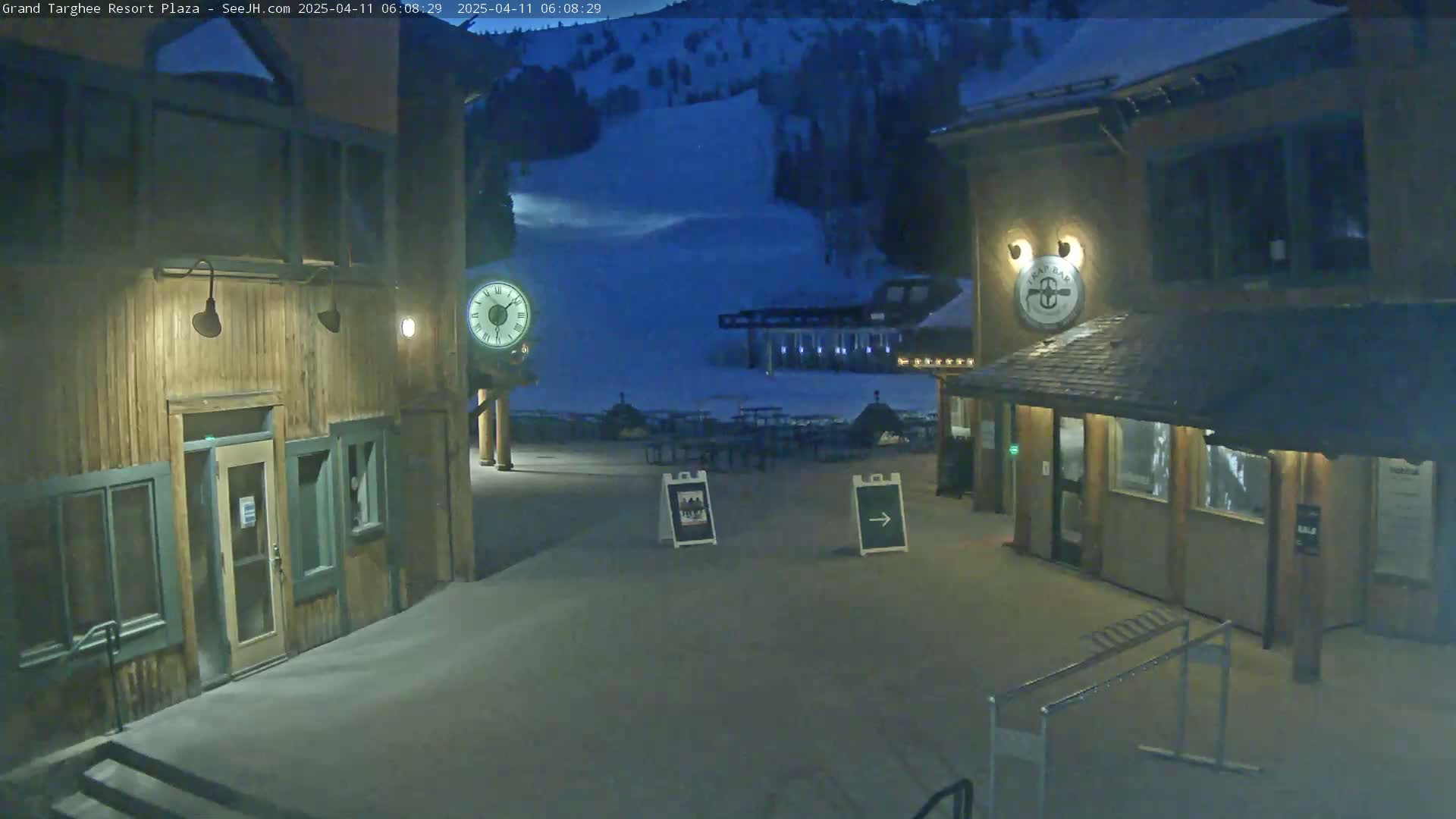 A snow-covered plaza at night is situated between two wooden buildings, one of which has a large clock on its exterior, with a ski slope visible in the background under a dark blue sky.