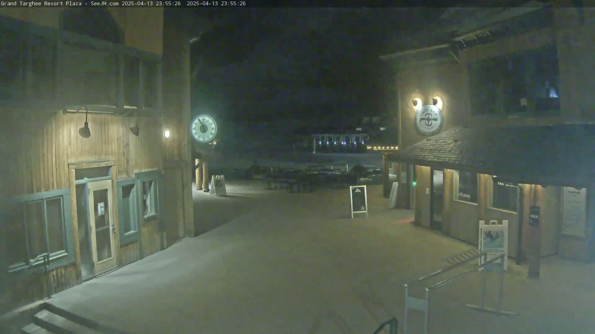 A nighttime view of a snow-covered plaza with two wooden buildings, one with a clock, under dark, clear skies.