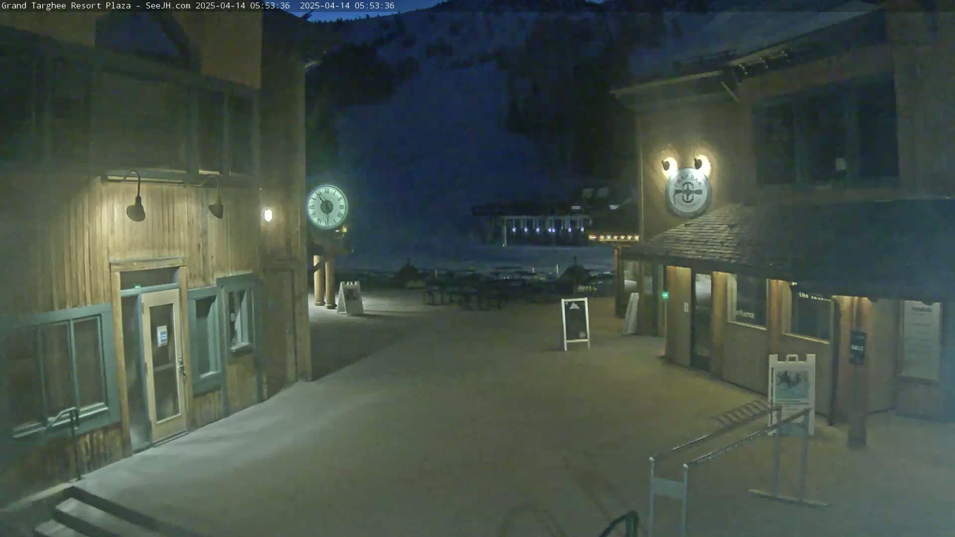 A nighttime view of a plaza with two wooden buildings, a clock, and outdoor seating against a dark mountainside.