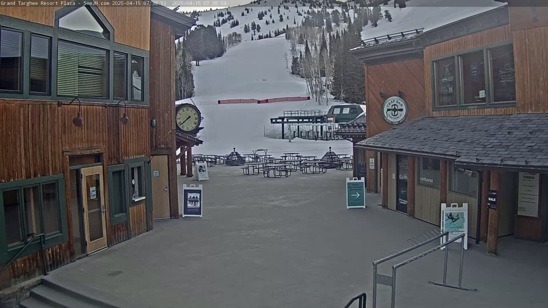 A plaza with several empty picnic tables sits between two wooden buildings overlooking a snow-covered ski slope on an overcast day.