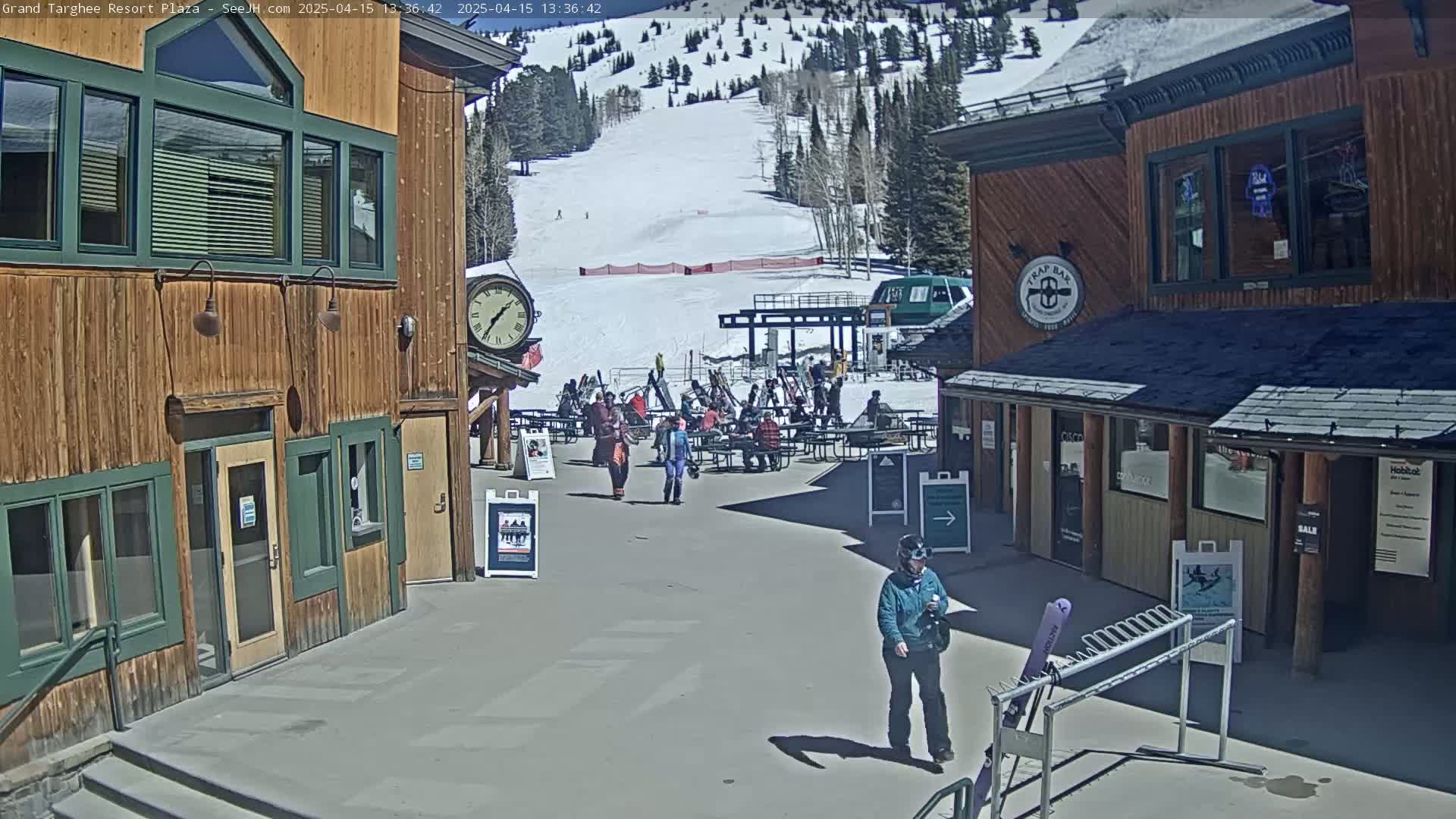 A sunny plaza with wooden buildings, a ski lift, and several people sitting at tables and walking around.
