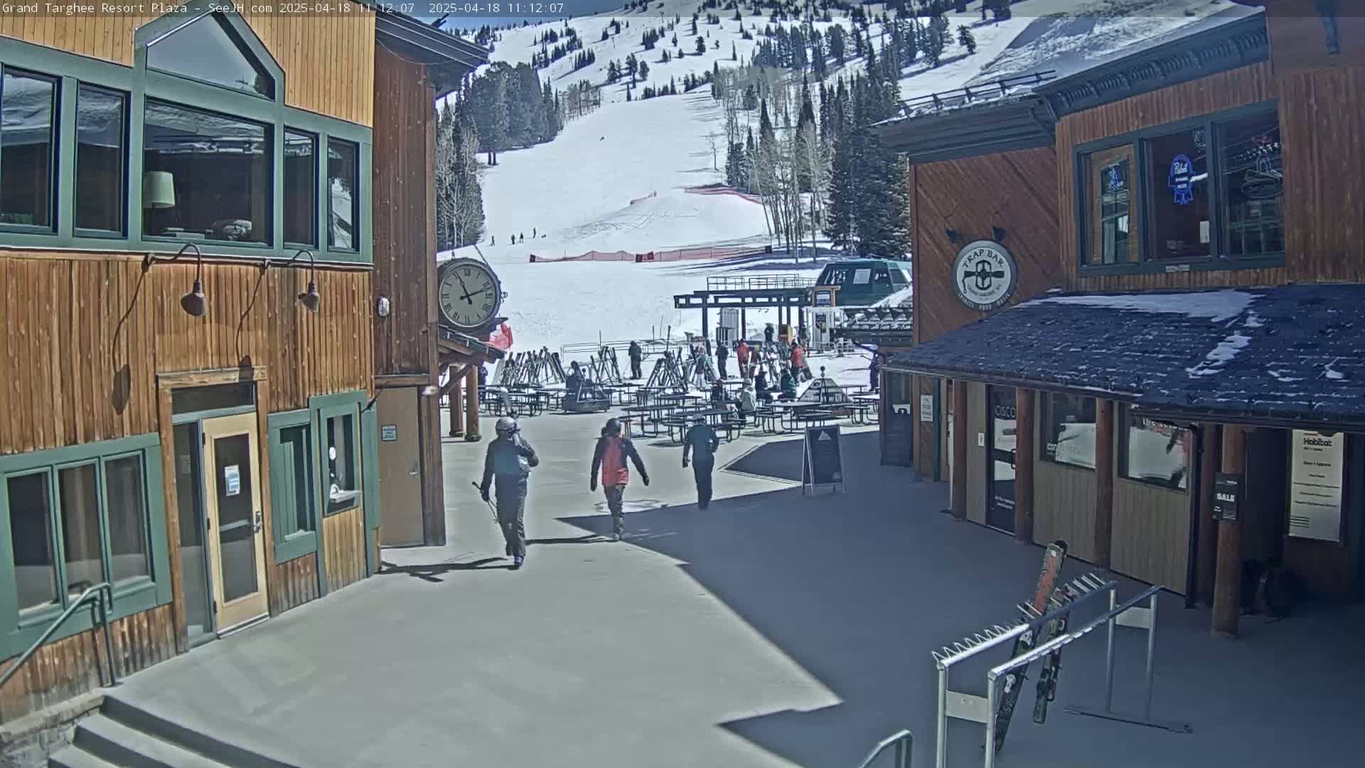 A sunny plaza at a ski resort shows several people walking past wooden buildings, a clock, picnic tables, and a ski lift in the background.