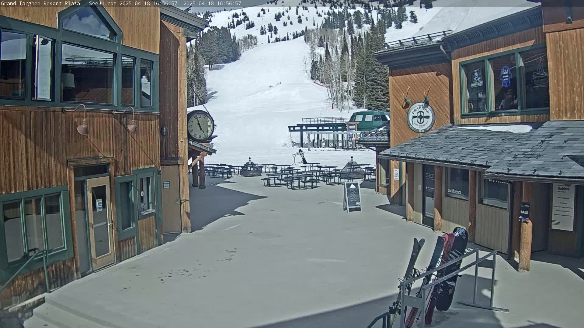 A plaza with wooden buildings, outdoor seating, a ski lift in the background, and a sunny day.