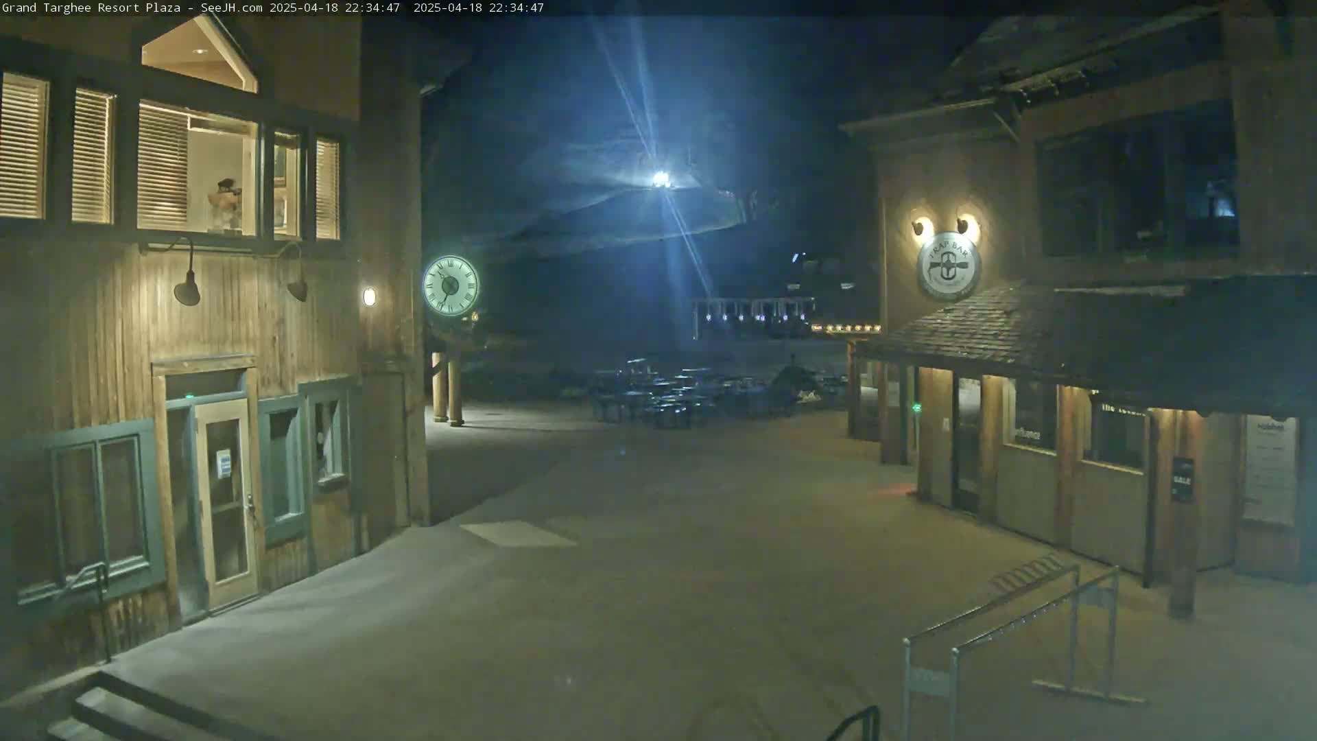 A nighttime view of a snow-covered plaza with two wooden buildings, one featuring a large clock, and outdoor seating in the center.