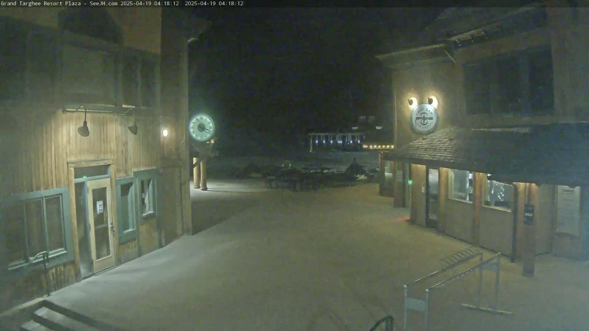 A snow-covered plaza at night, illuminated by lights on wooden buildings, with a large clock visible in the center.
