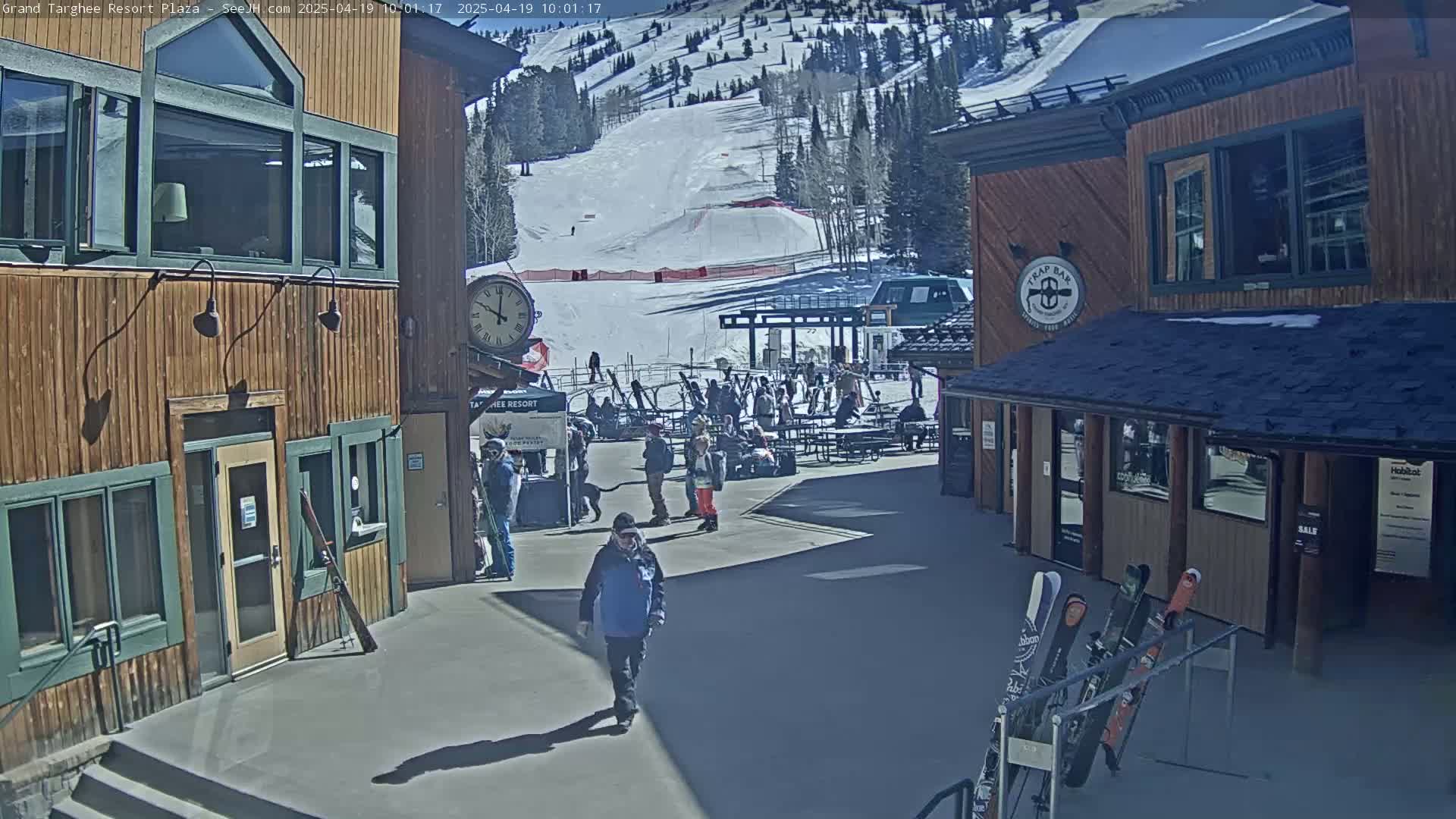 A sunny plaza at a ski resort with several buildings, people milling around, and skis leaning against a rack, with a snowy mountainside visible in the background.
