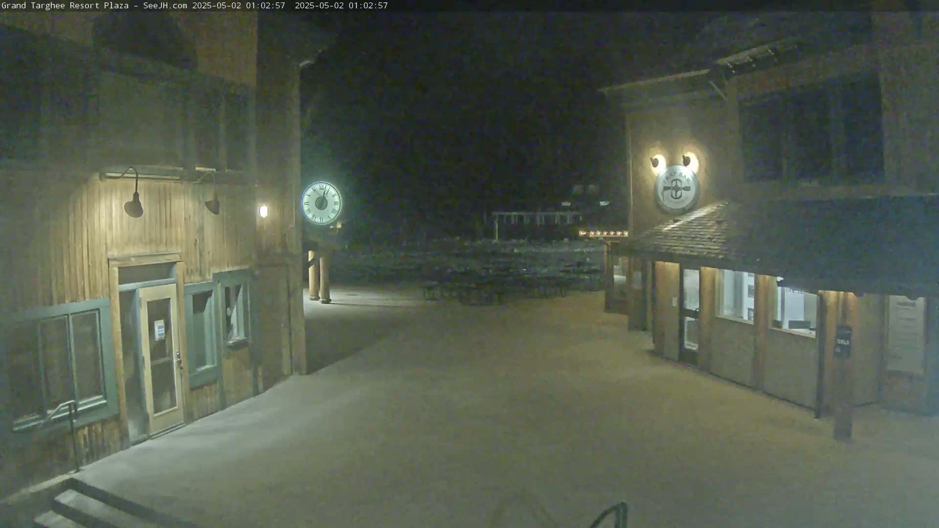 A snow-covered plaza at night is illuminated, showing two wooden buildings with clocks, and a sparsely populated seating area in between.