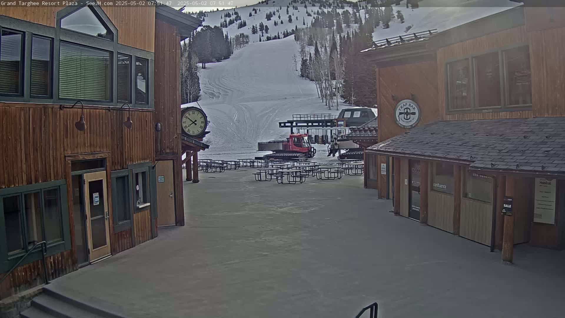 A plaza with wooden buildings, a snow-covered ski slope in the background, and several empty picnic tables sits under an overcast sky.
