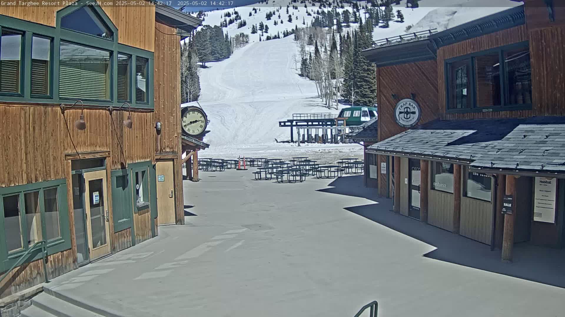 A plaza with wooden buildings and empty picnic tables overlooks a snow-covered ski slope on a sunny day.