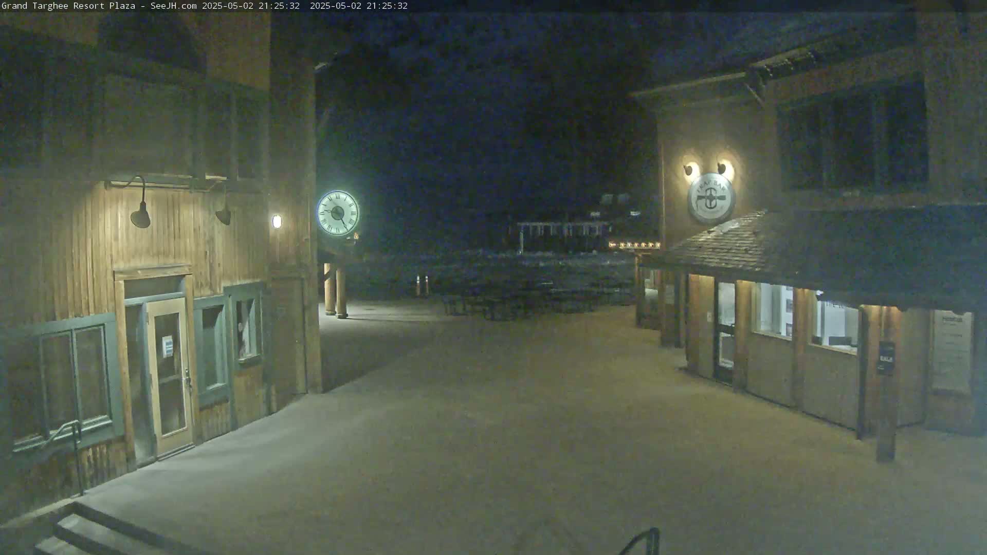 A snow-covered plaza at night, illuminated by lights on wooden buildings, features a large clock and outdoor seating.