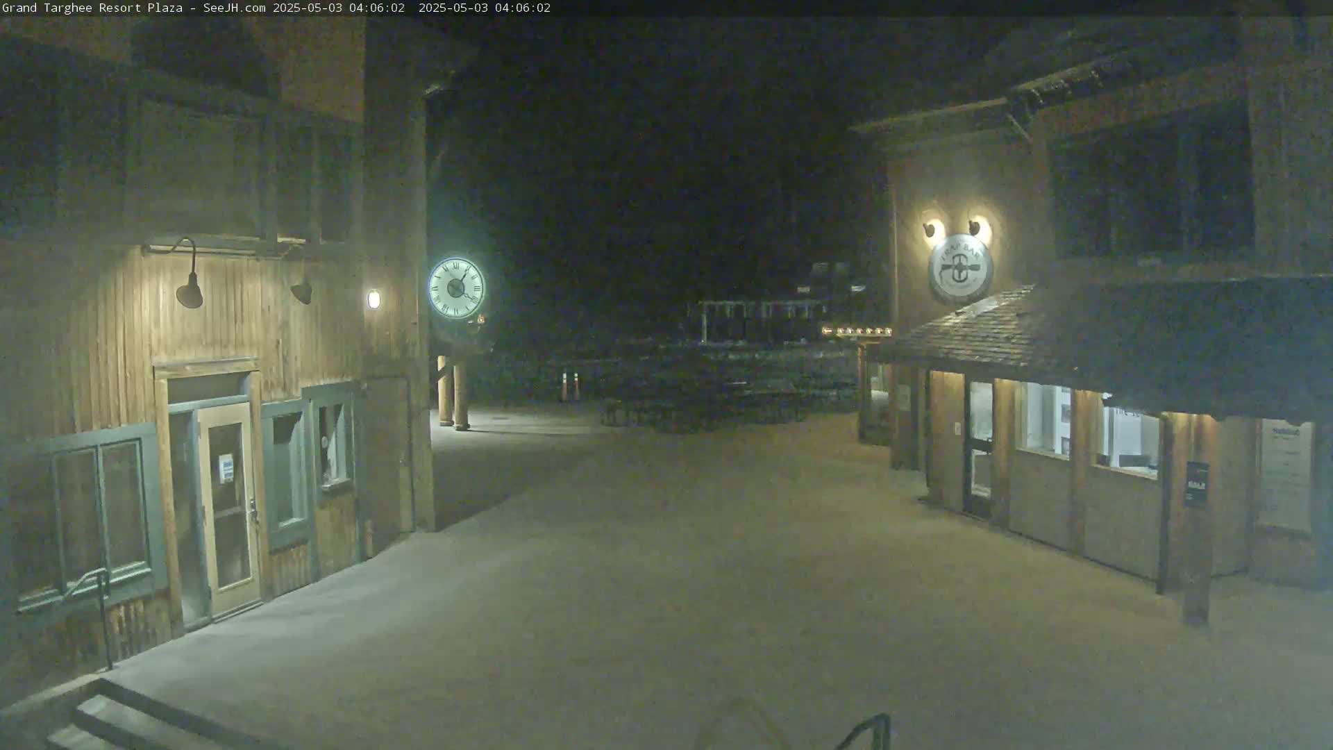 A plaza at night, illuminated by artificial light, shows two wooden buildings with a snow-covered ground.