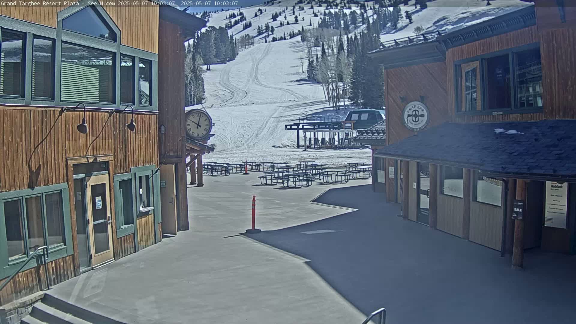 A plaza with wooden buildings, numerous picnic tables, a ski slope with a chairlift in the background, and sunny weather.