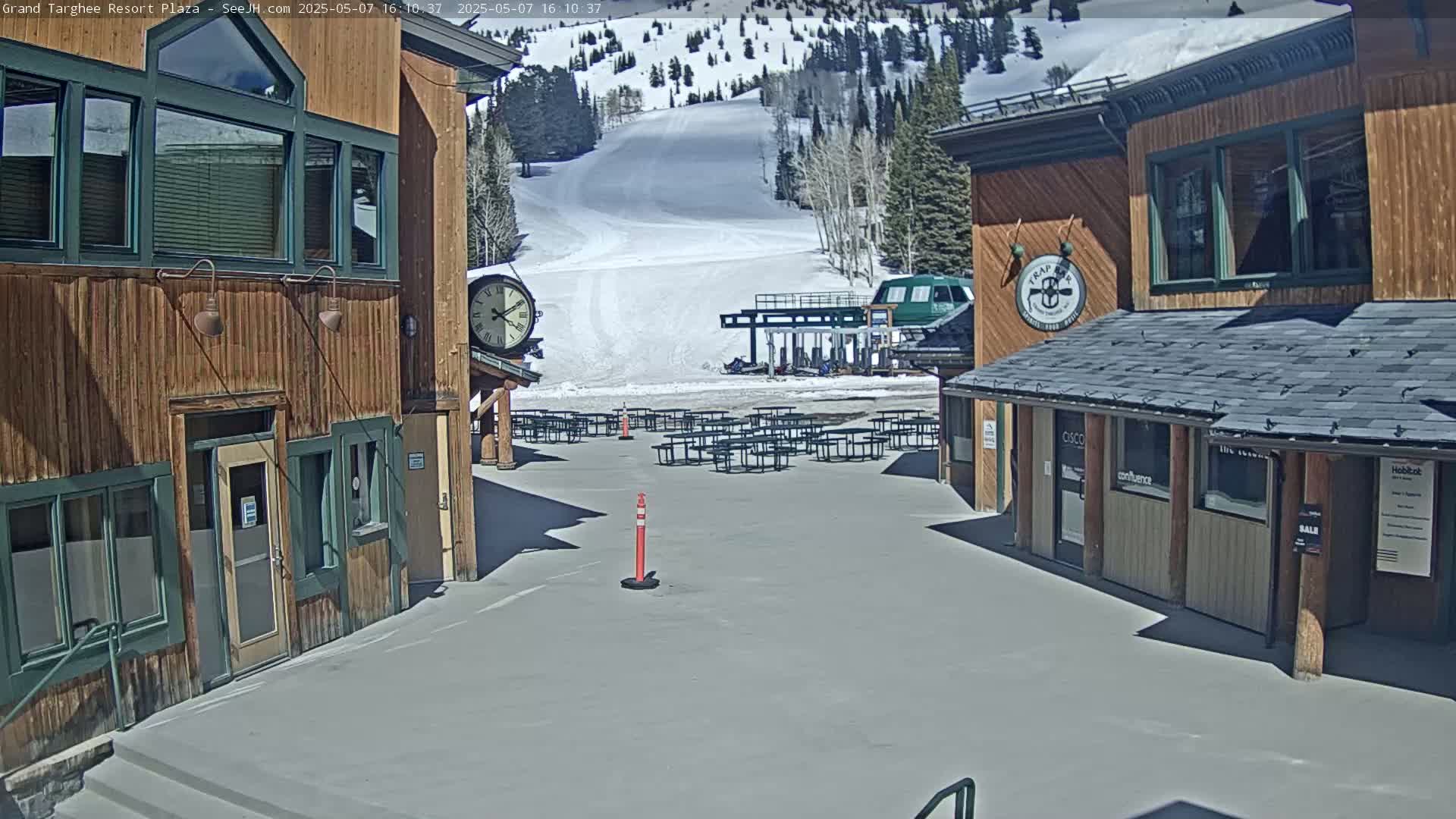 A plaza with wooden buildings, numerous empty picnic tables, a ski lift, and a snowy mountainside in the background under sunny conditions.