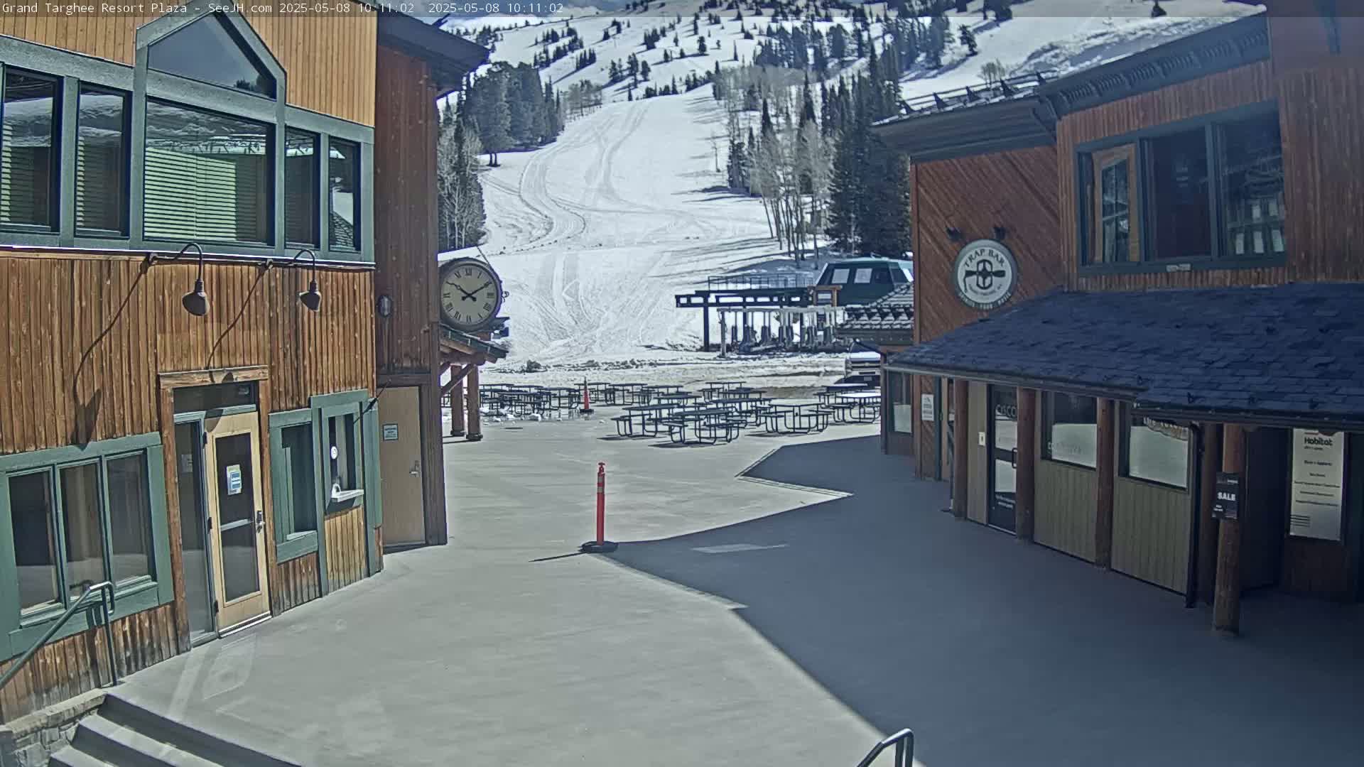 A plaza with numerous empty picnic tables sits between two wood-sided buildings, overlooking a snow-covered ski slope on a sunny day.