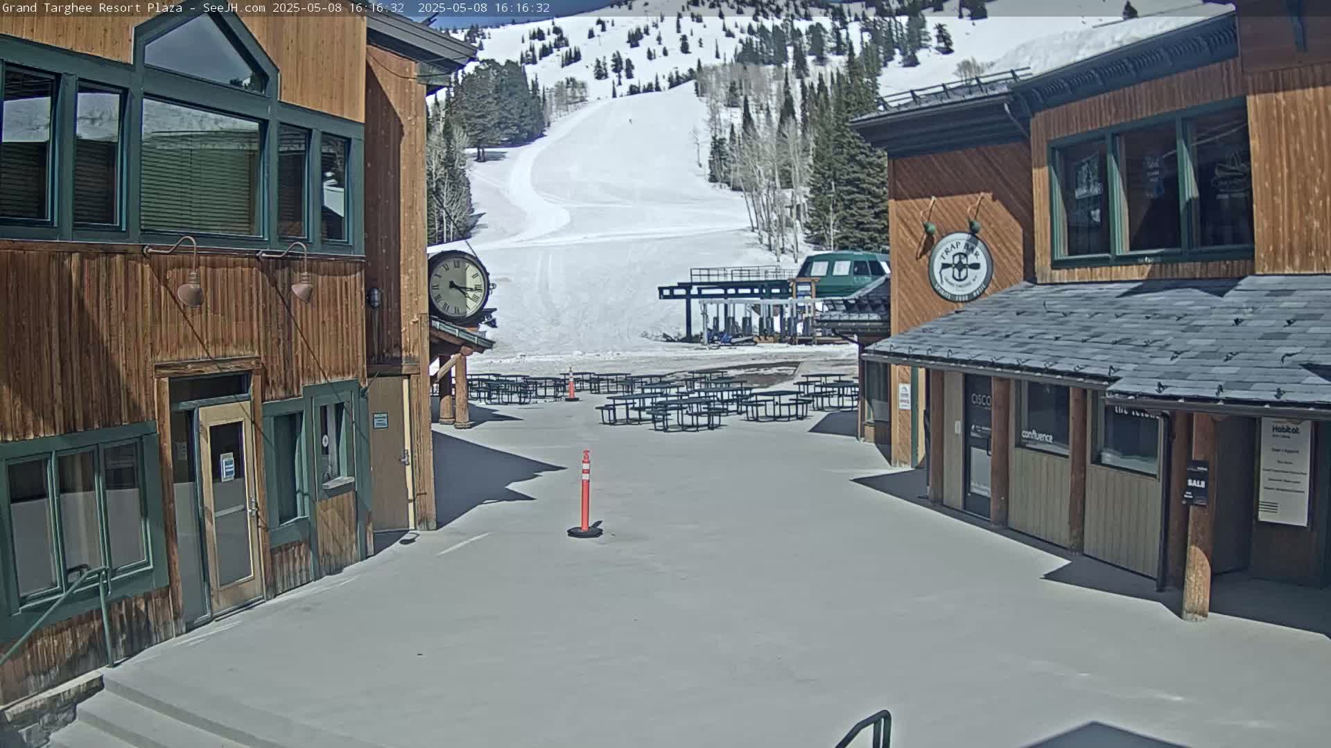 A plaza with wooden buildings, empty picnic tables, and a ski lift visible in the background on a sunny day.