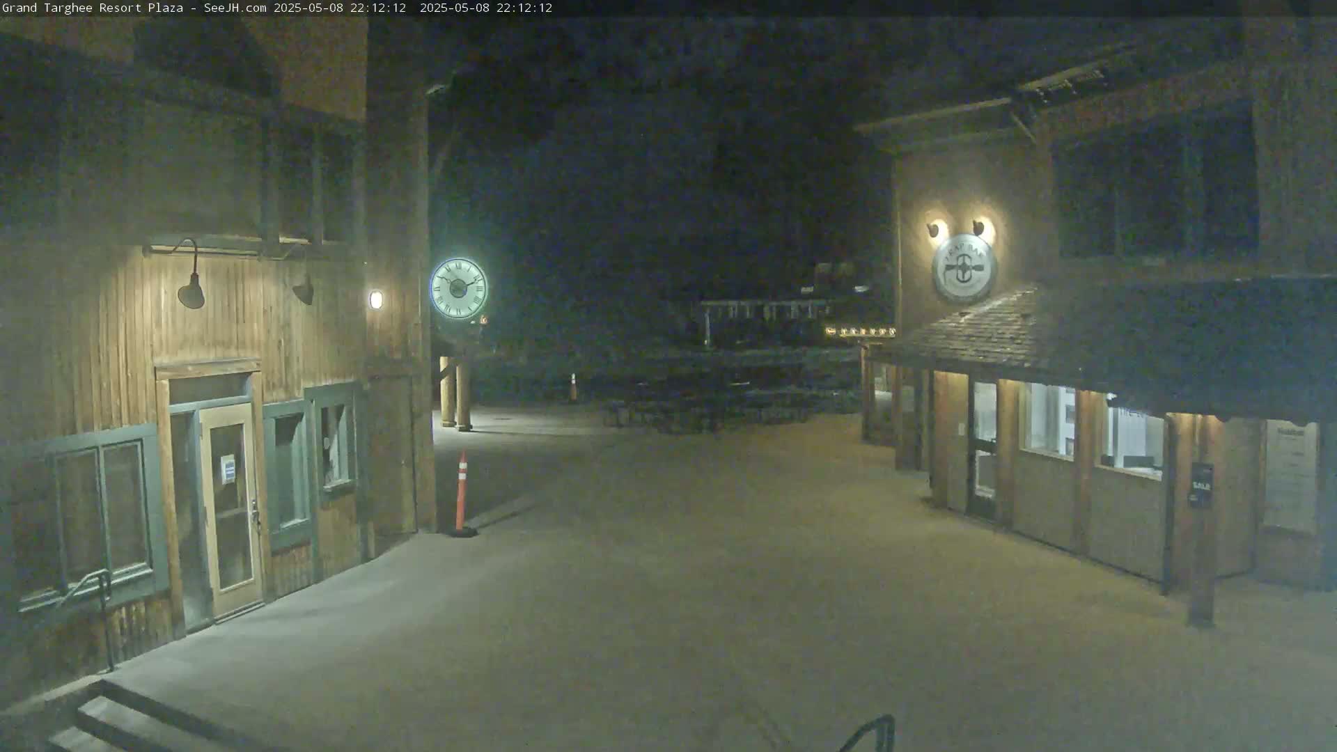 A nighttime, snow-covered plaza with two wooden buildings, one featuring a clock, is illuminated by artificial light.