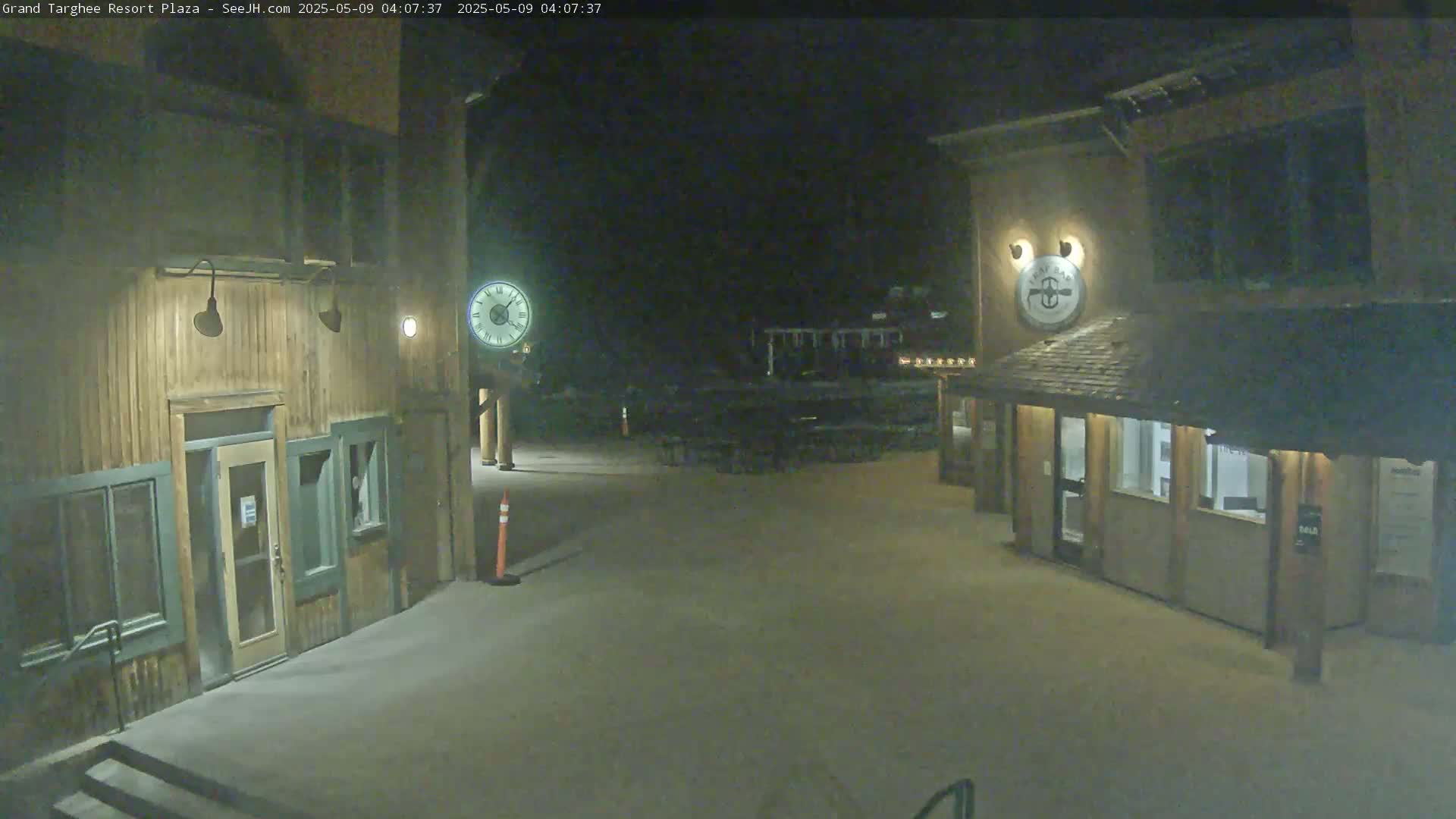 A snow-covered plaza at night, illuminated by artificial light, is flanked by two wooden buildings with entrances and windows.