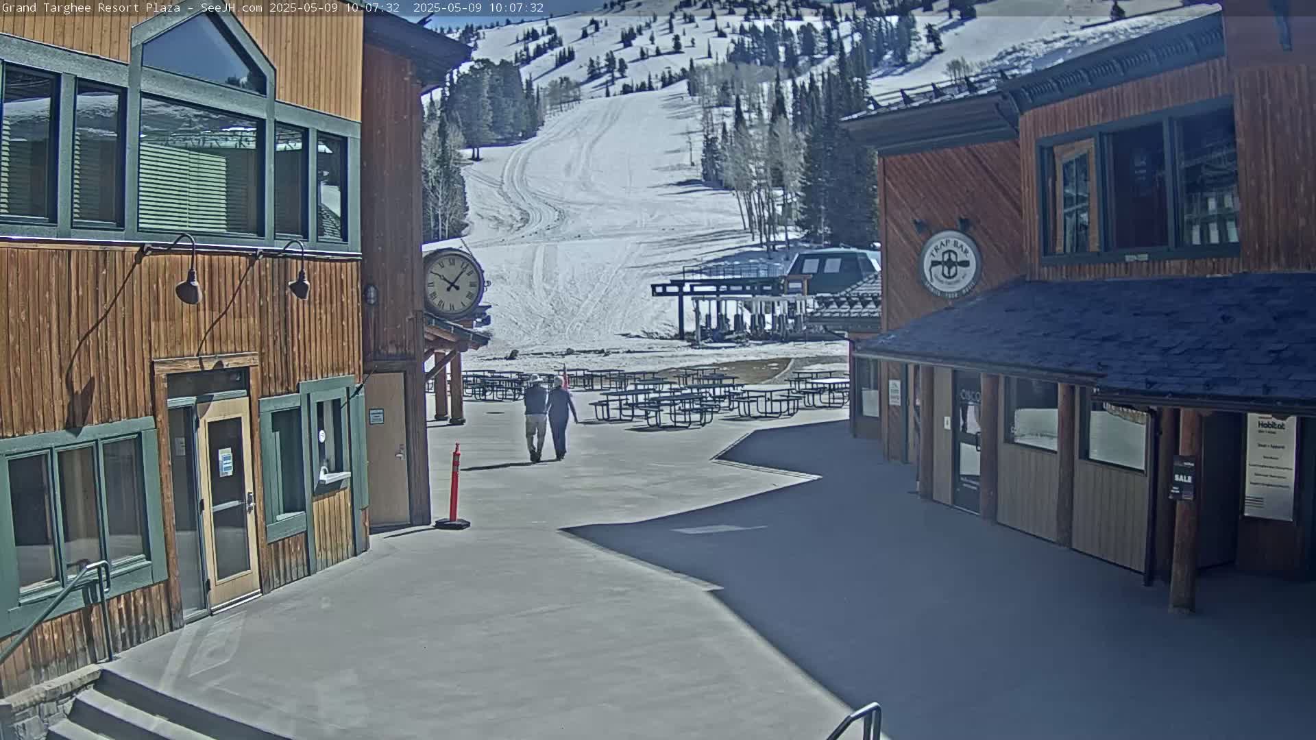 Two people walk toward a plaza with wooden buildings, picnic tables, and a ski slope visible in the background under a sunny sky.