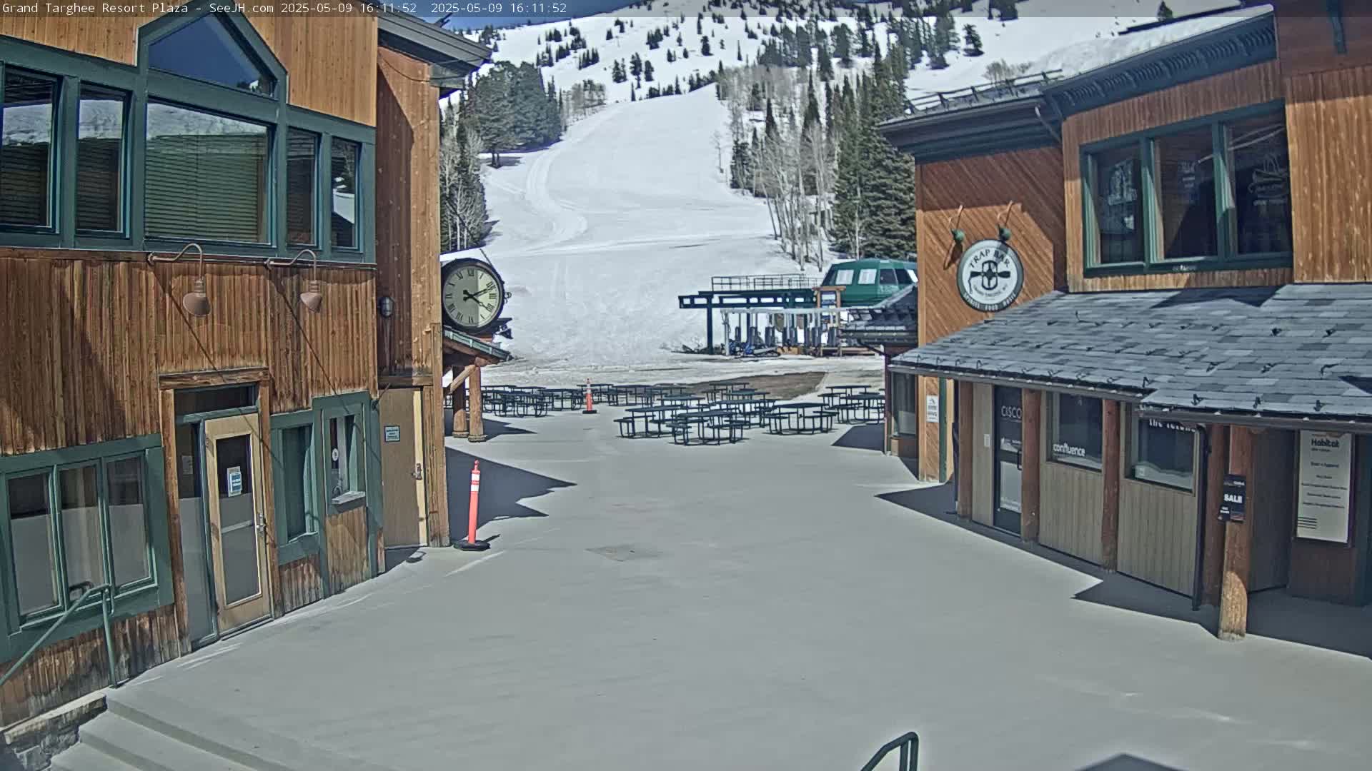 A plaza with several picnic tables sits between two wooden buildings under a sunny sky, overlooking a snow-covered ski slope with a chairlift.