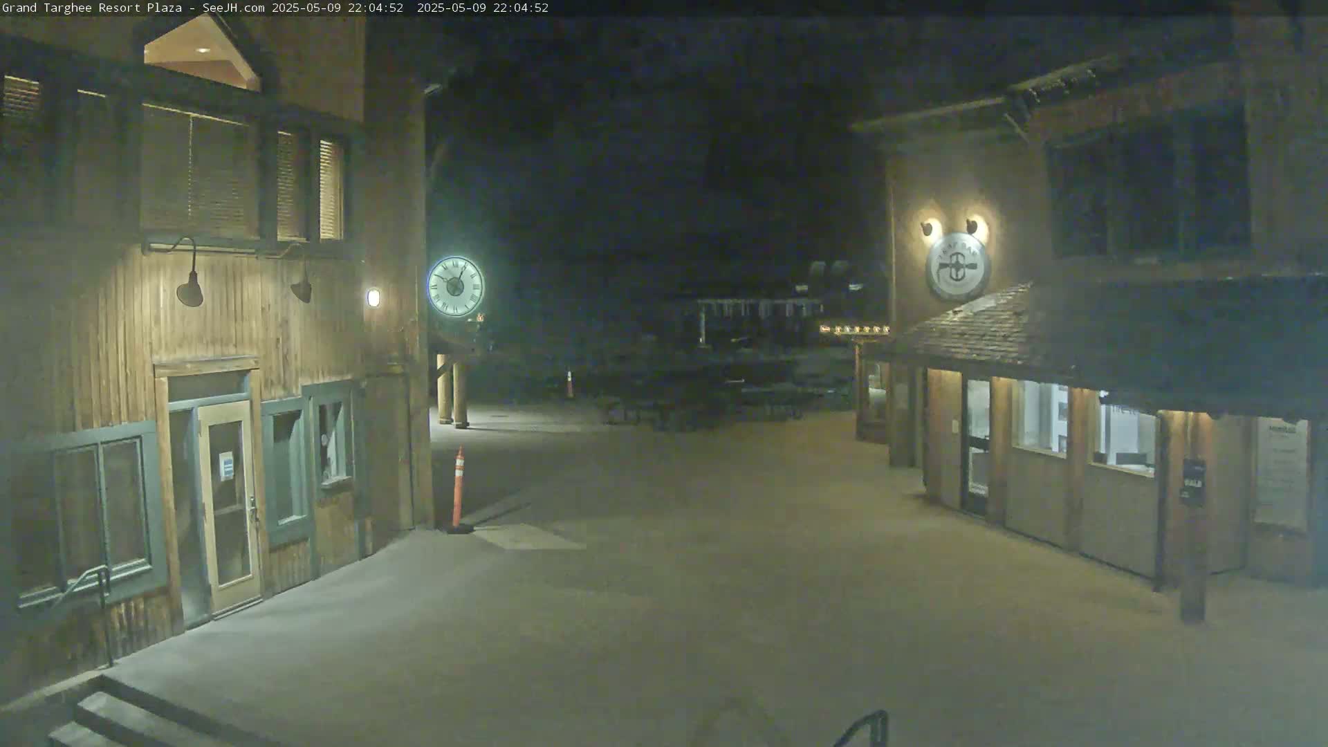 At night, two wooden buildings with lights on are visible in a snow-covered plaza.
