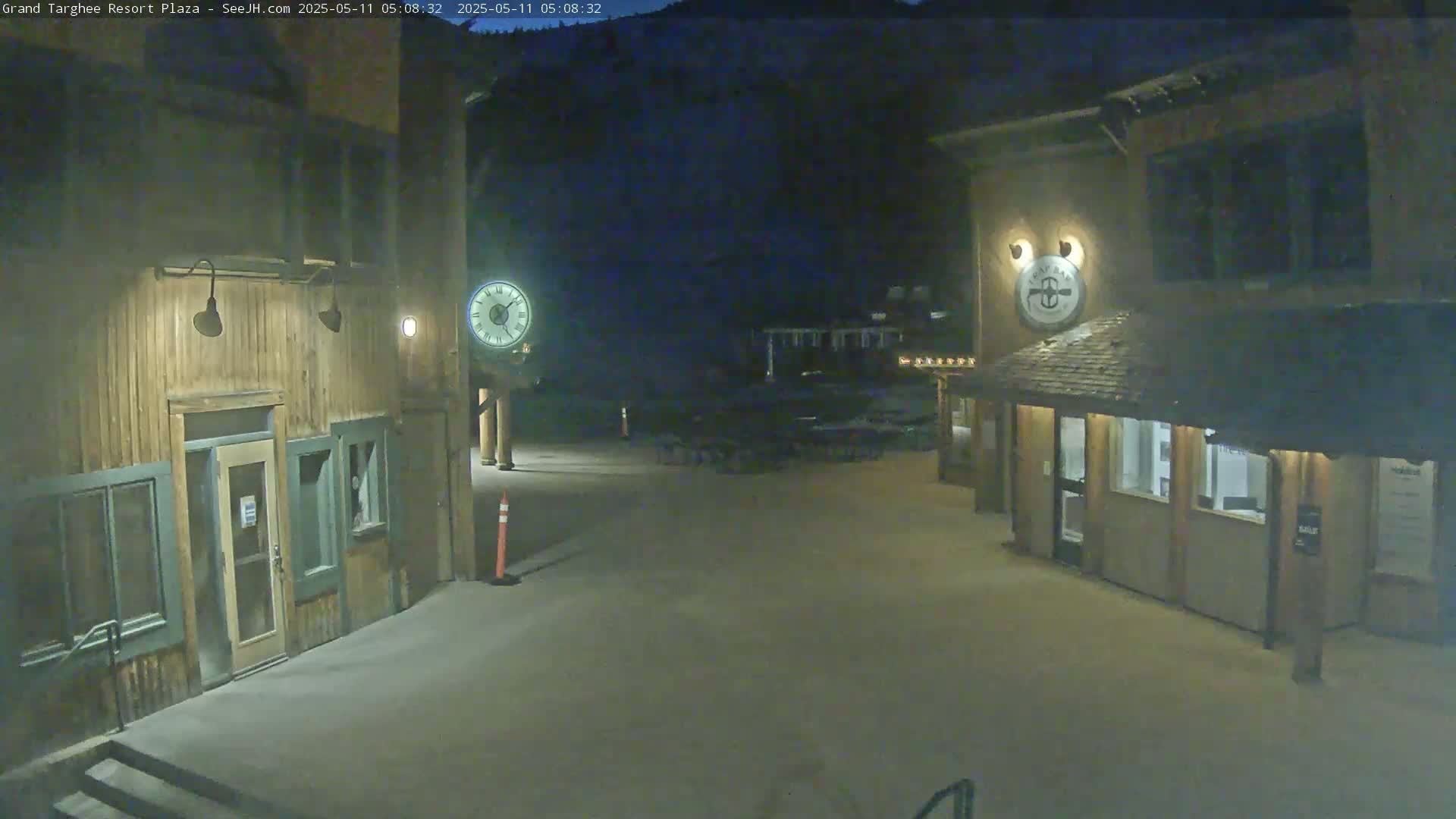 A plaza at night, illuminated by artificial light, features two wooden buildings with clocks, and a dark mountainside in the background.