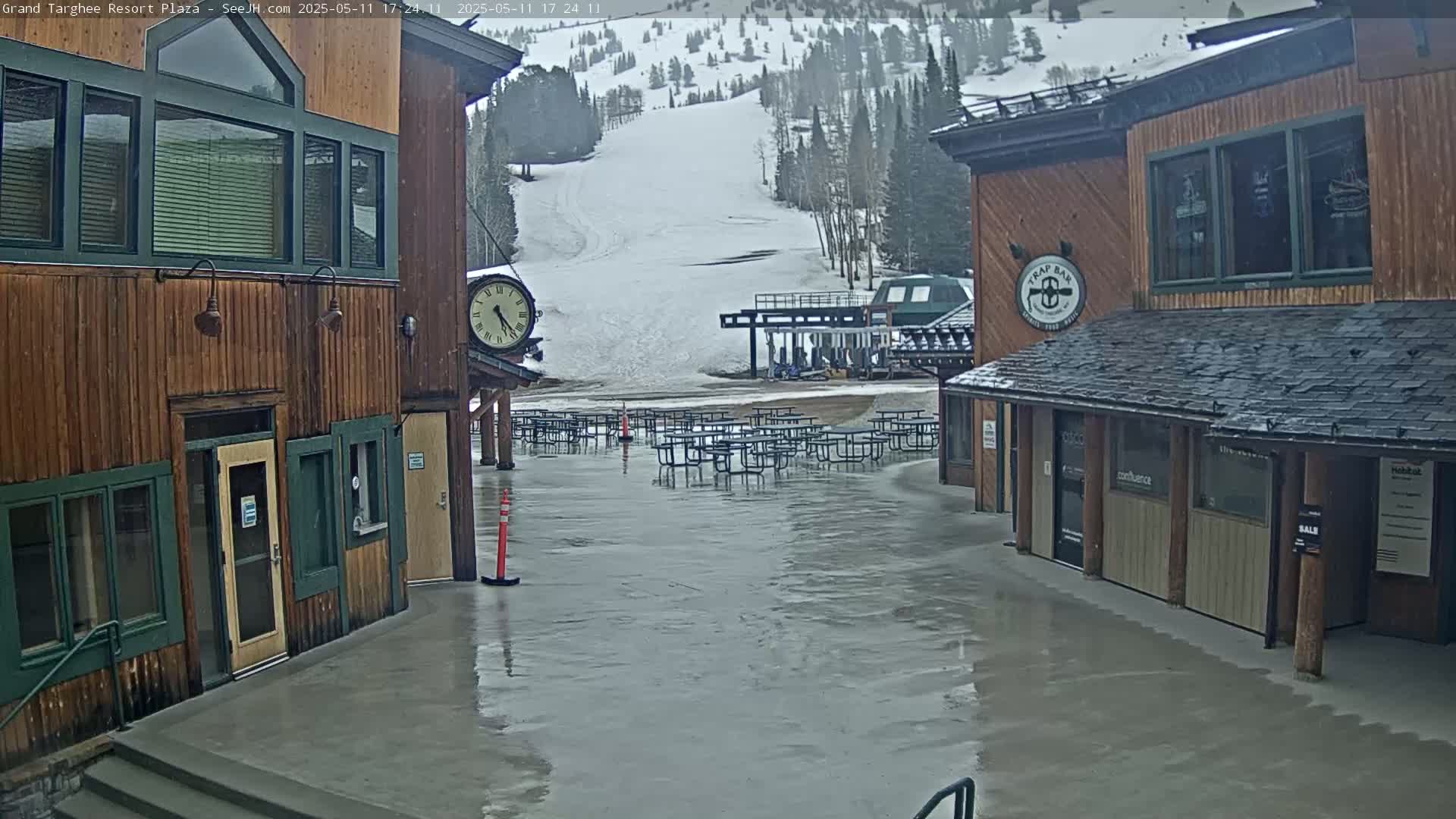 A wet plaza with several empty picnic tables sits between two wooden buildings overlooking a snowy ski slope.