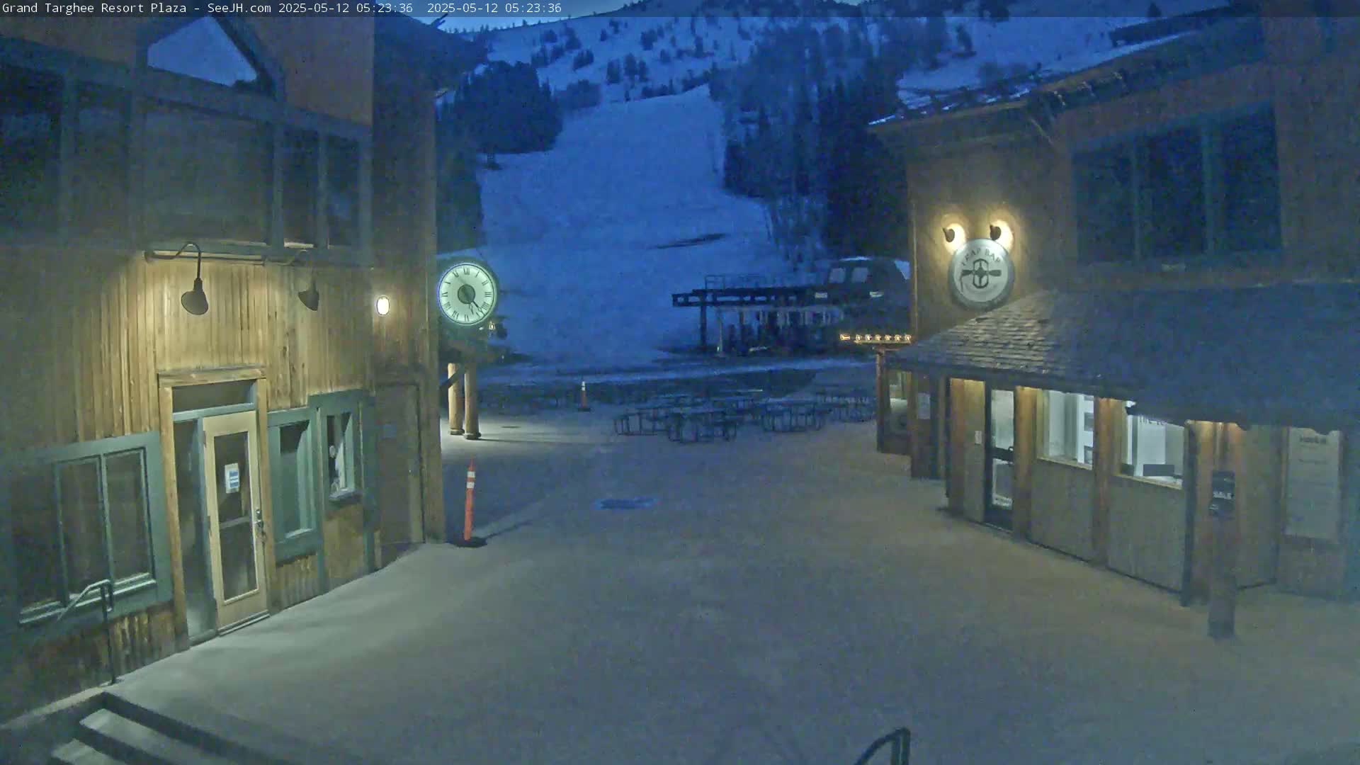 A snow-covered plaza at night with two wooden buildings, a ski lift, and picnic tables, under a dark sky.