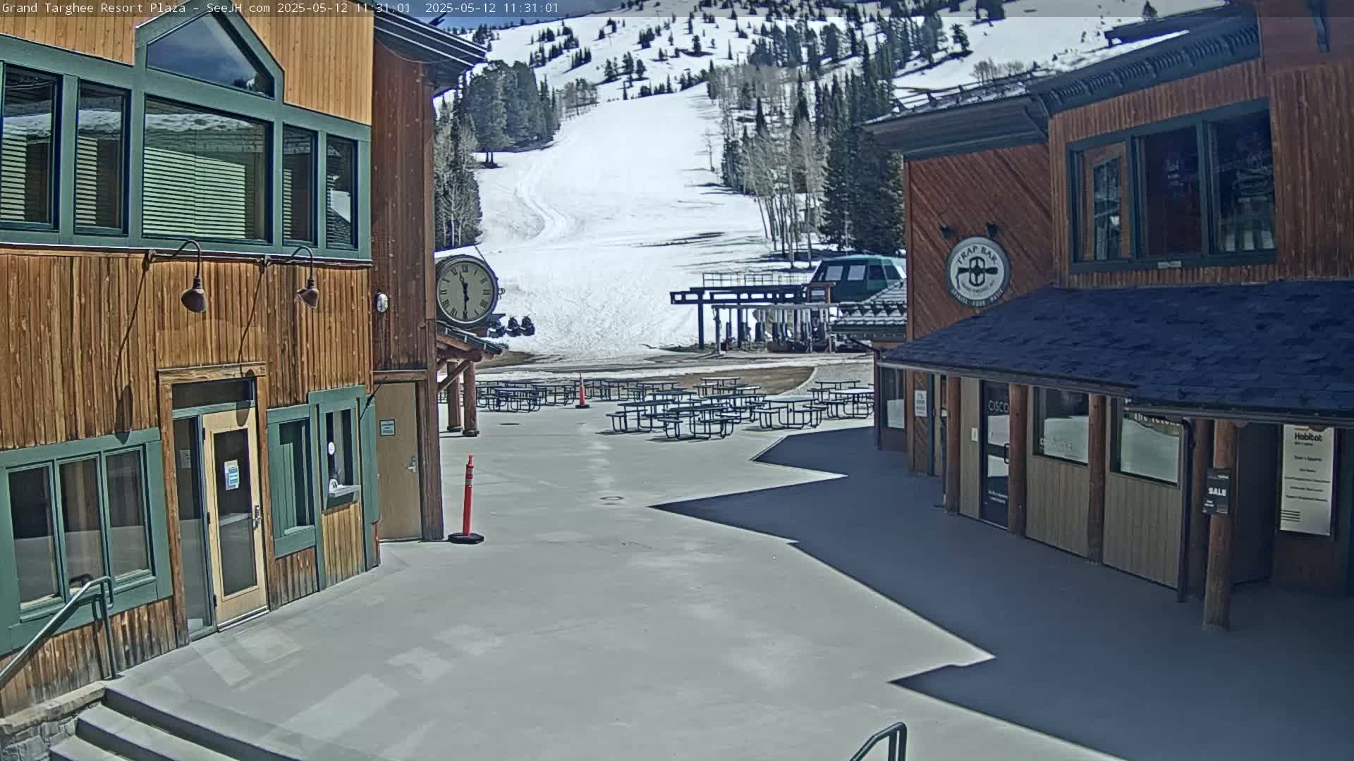 A plaza with several picnic tables sits between two wooden buildings overlooking a snow-covered ski slope on a sunny day.