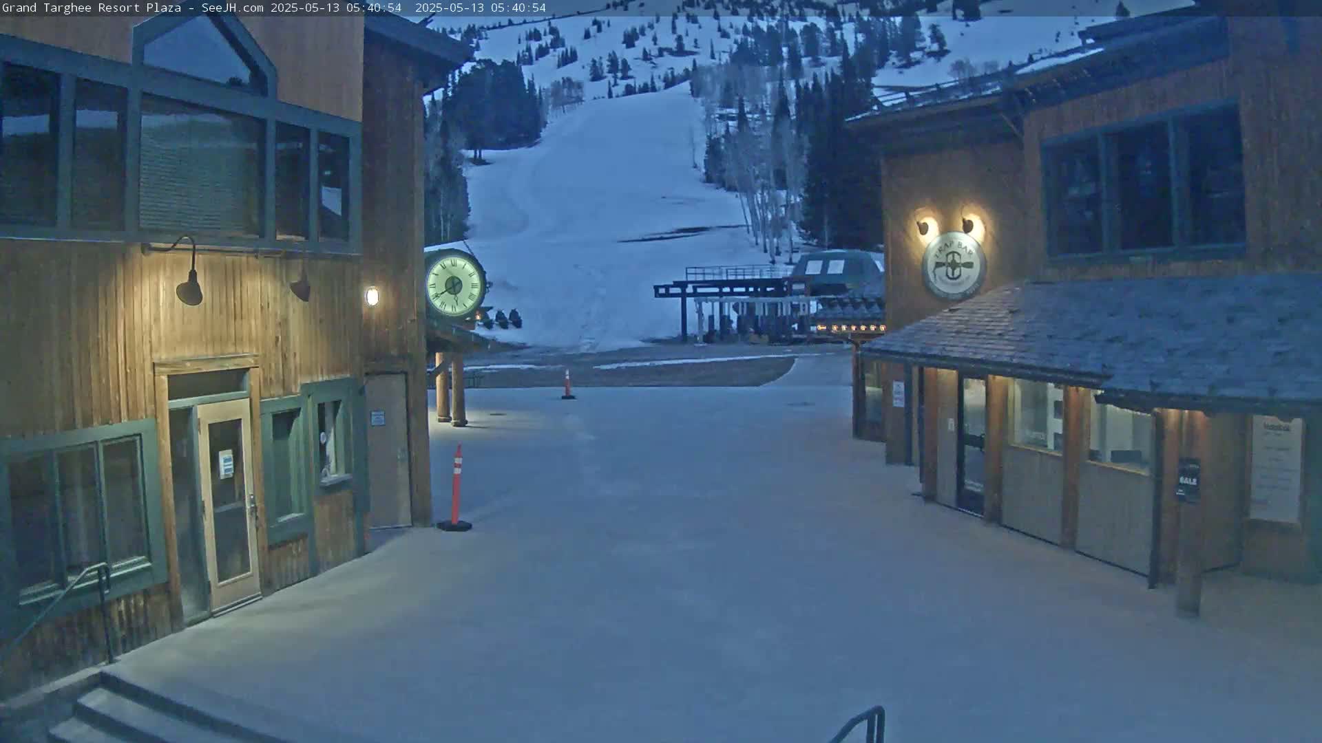 A snow-covered plaza with two wooden buildings, one with a clock, overlooks a ski slope under twilight conditions.
