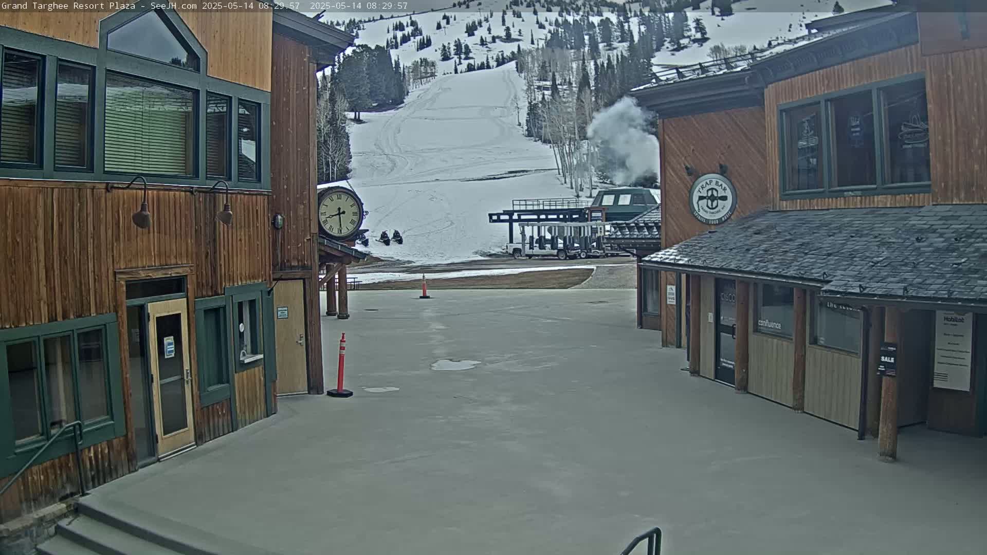 A plaza with two wooden buildings overlooks a snowy ski slope with a ski lift and some light smoke in the air.