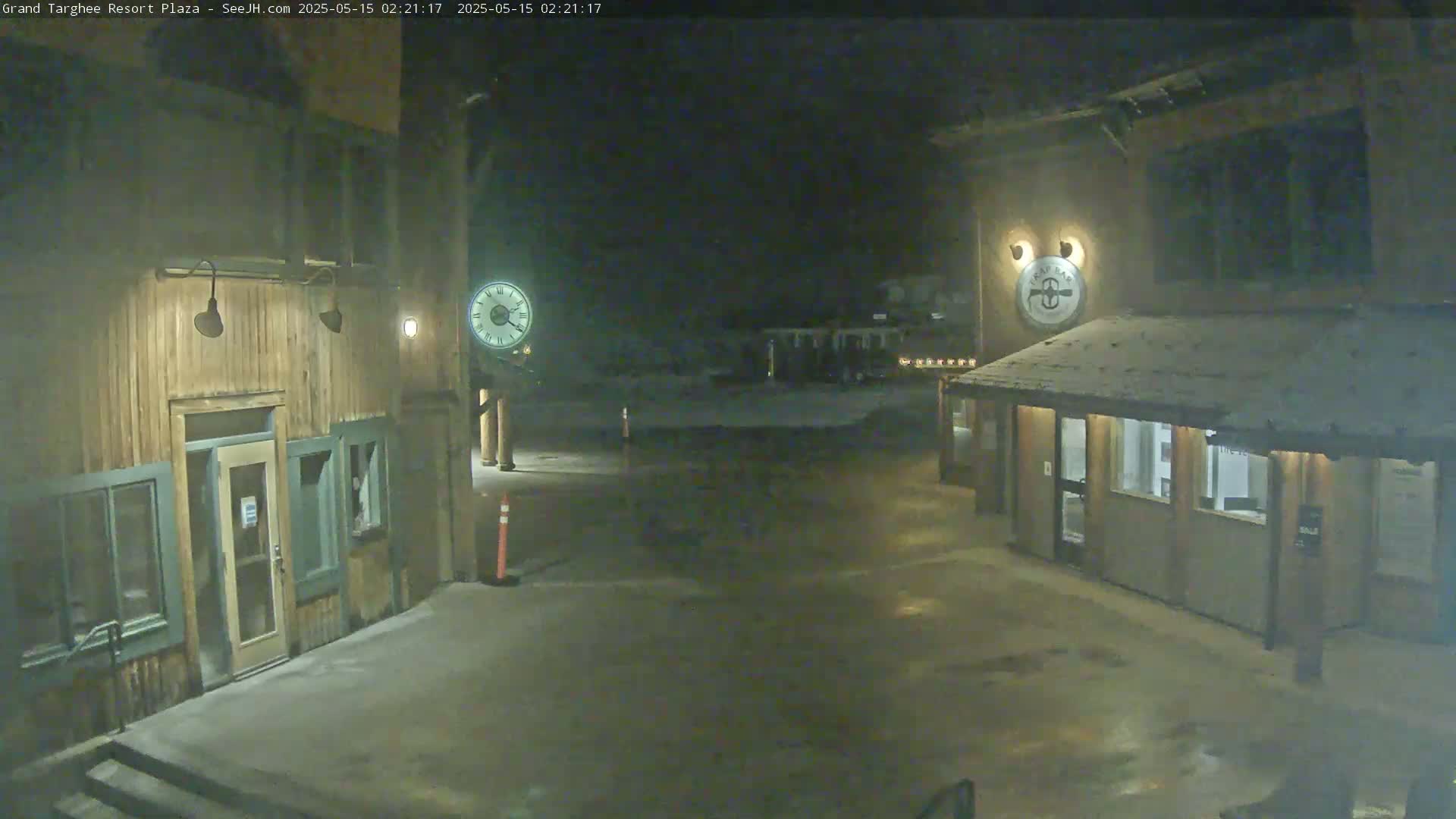 A nighttime, snow-dusted plaza with two wooden buildings, one displaying a clock and the other a circular emblem.