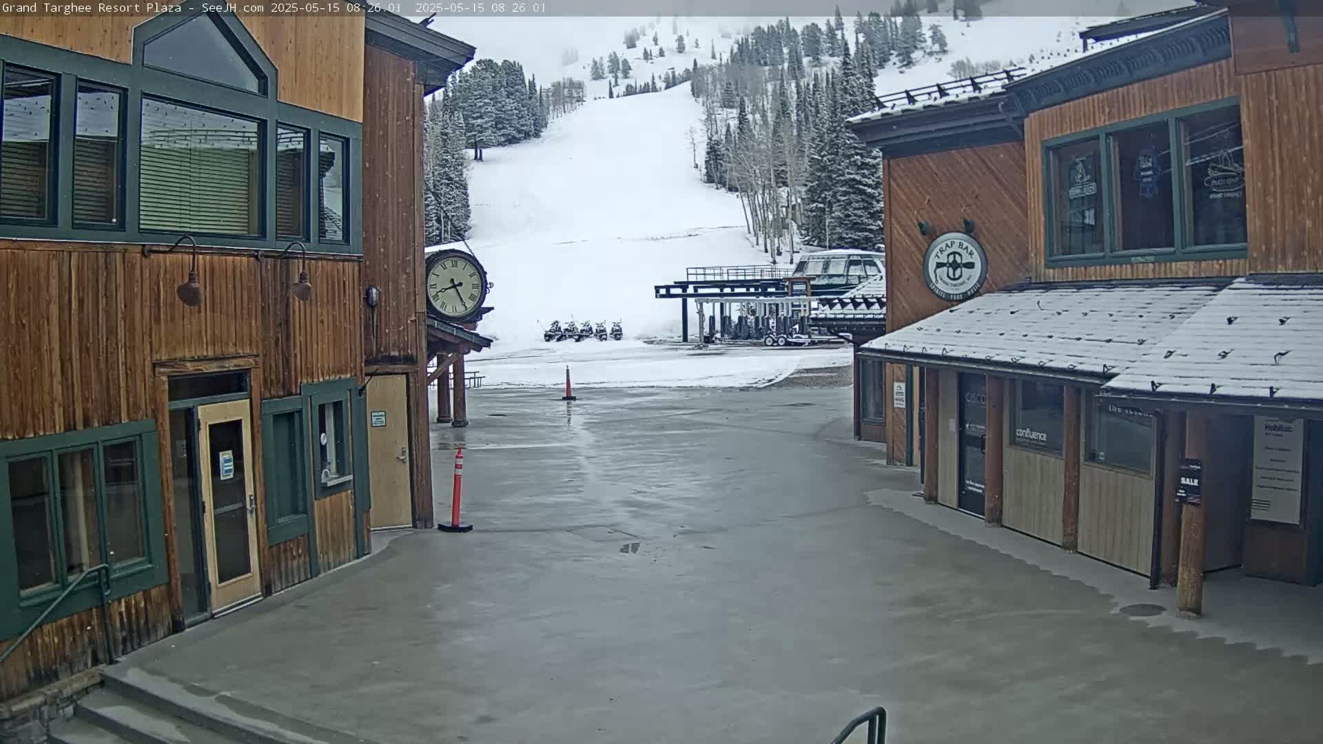 A plaza with wooden buildings, a snowy ski slope in the background, and light snow falling.