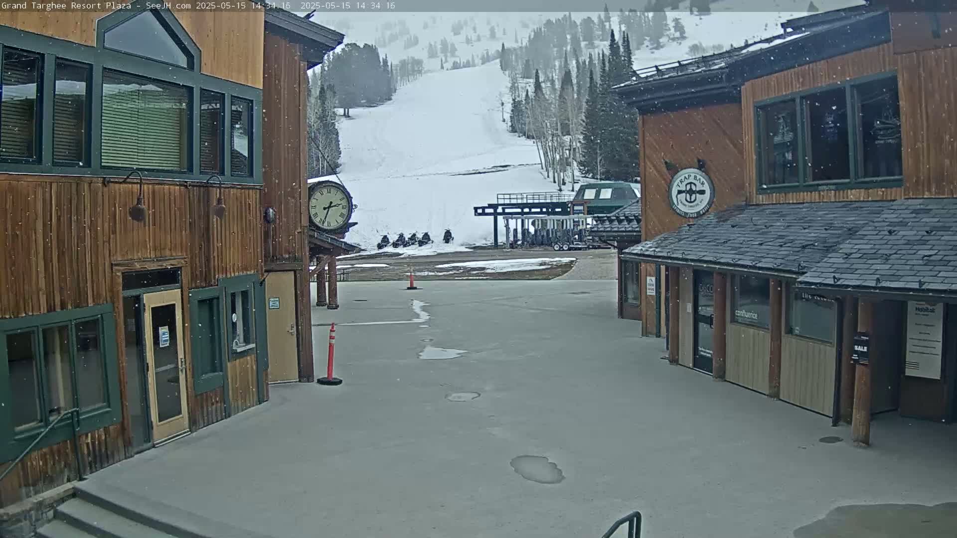 Snow falls on a plaza between two wooden buildings, overlooking a ski slope with snowmobiles parked near a ski lift.