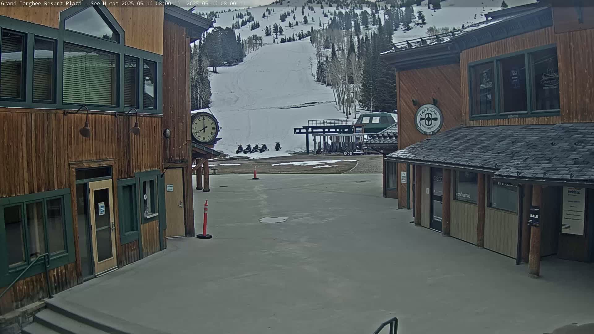 A plaza with two wooden buildings flanking a paved area, overlooking a snow-covered ski slope with a chairlift and snowmobiles parked nearby, under an overcast sky.