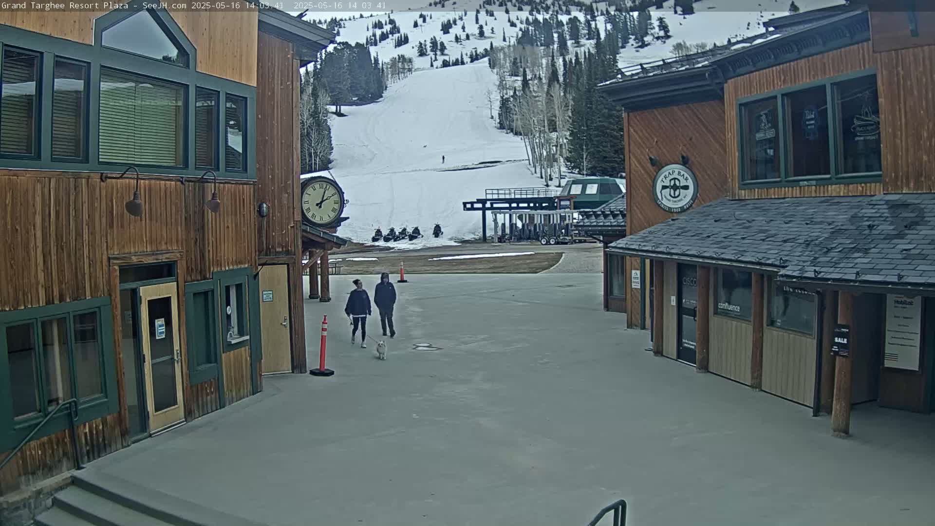 A couple walks a dog past two wooden buildings in a plaza overlooking a snow-covered ski slope on an overcast day.