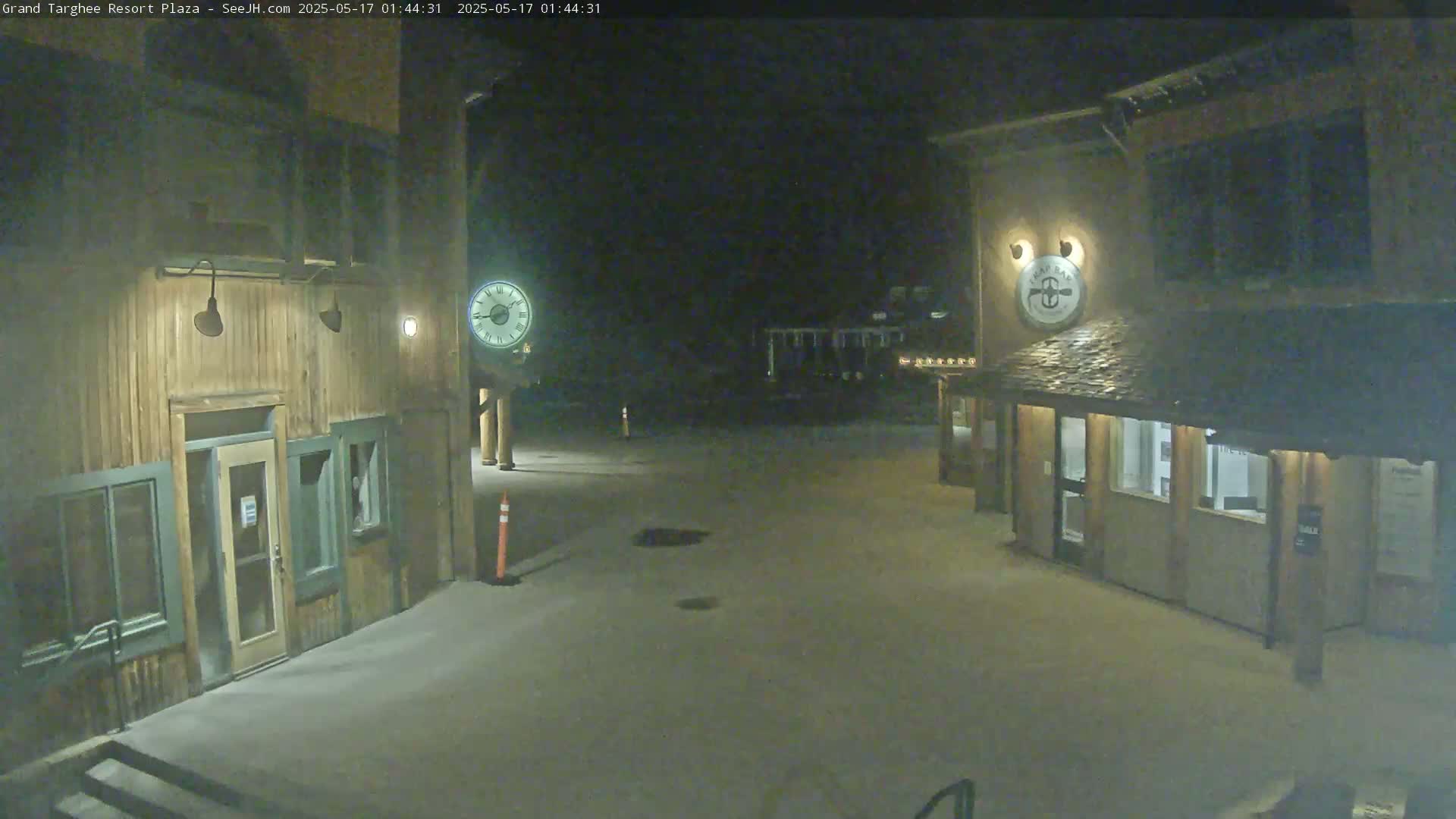 A snow-covered plaza at night is lit by artificial light, showing two wooden buildings with clocks on their facades.