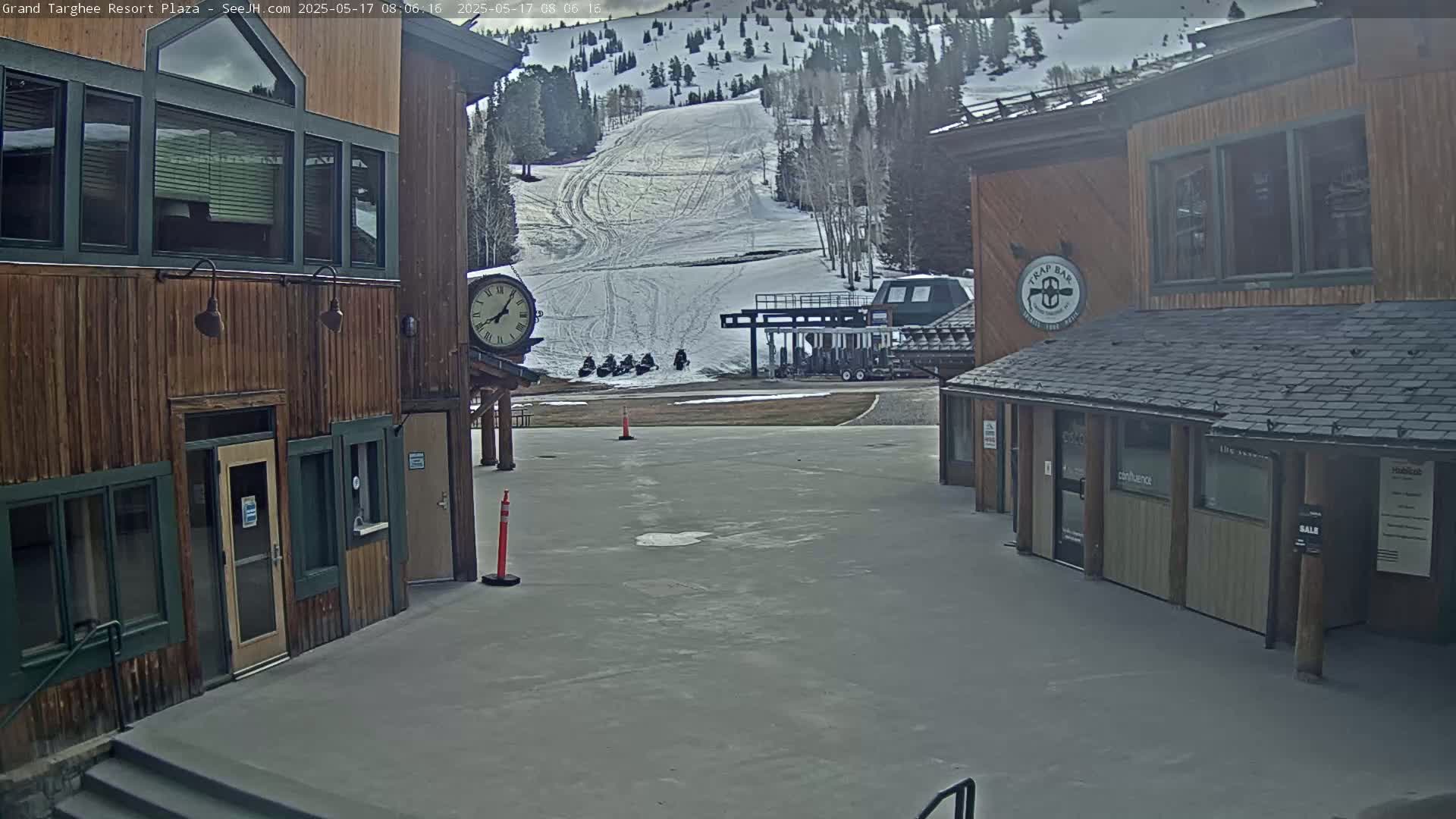A plaza with two wood-sided buildings overlooks a snow-covered ski slope with several snowmobiles parked near a ski lift on a partly cloudy day.