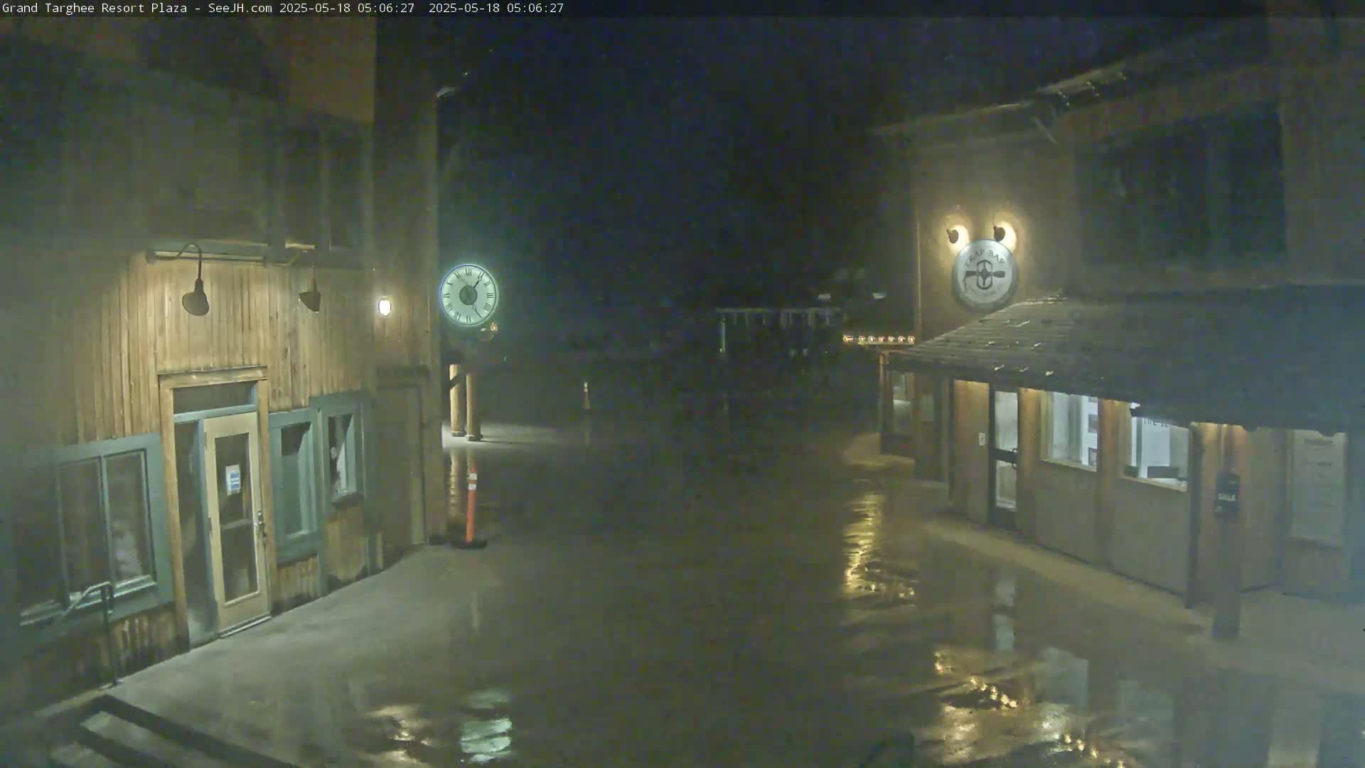 A wet plaza at night, illuminated by lights on two wooden buildings, one of which displays a clock.