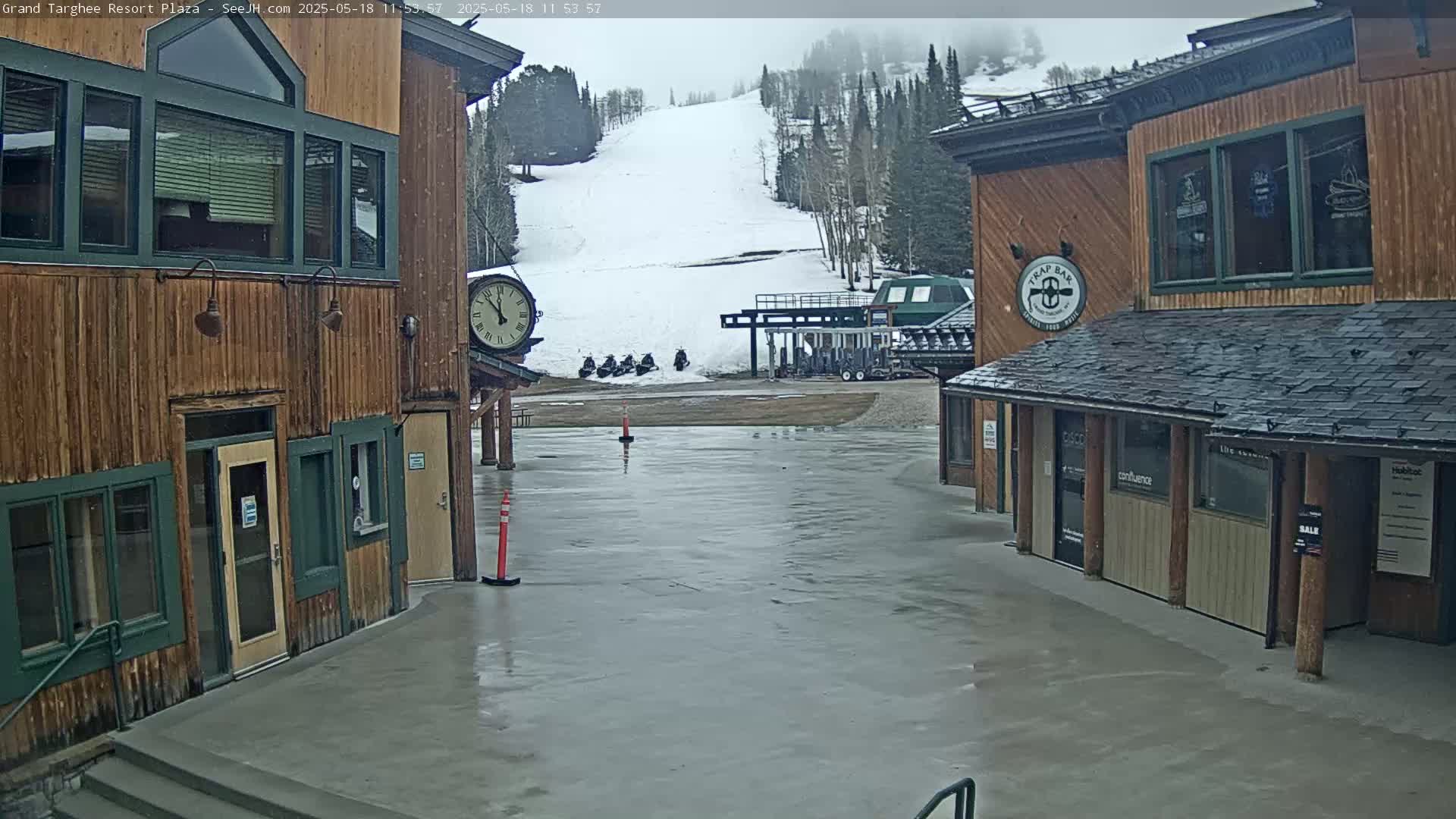 A wet plaza between two wooden buildings overlooks a snow-covered ski slope with several snowmobiles parked near a ski lift.
