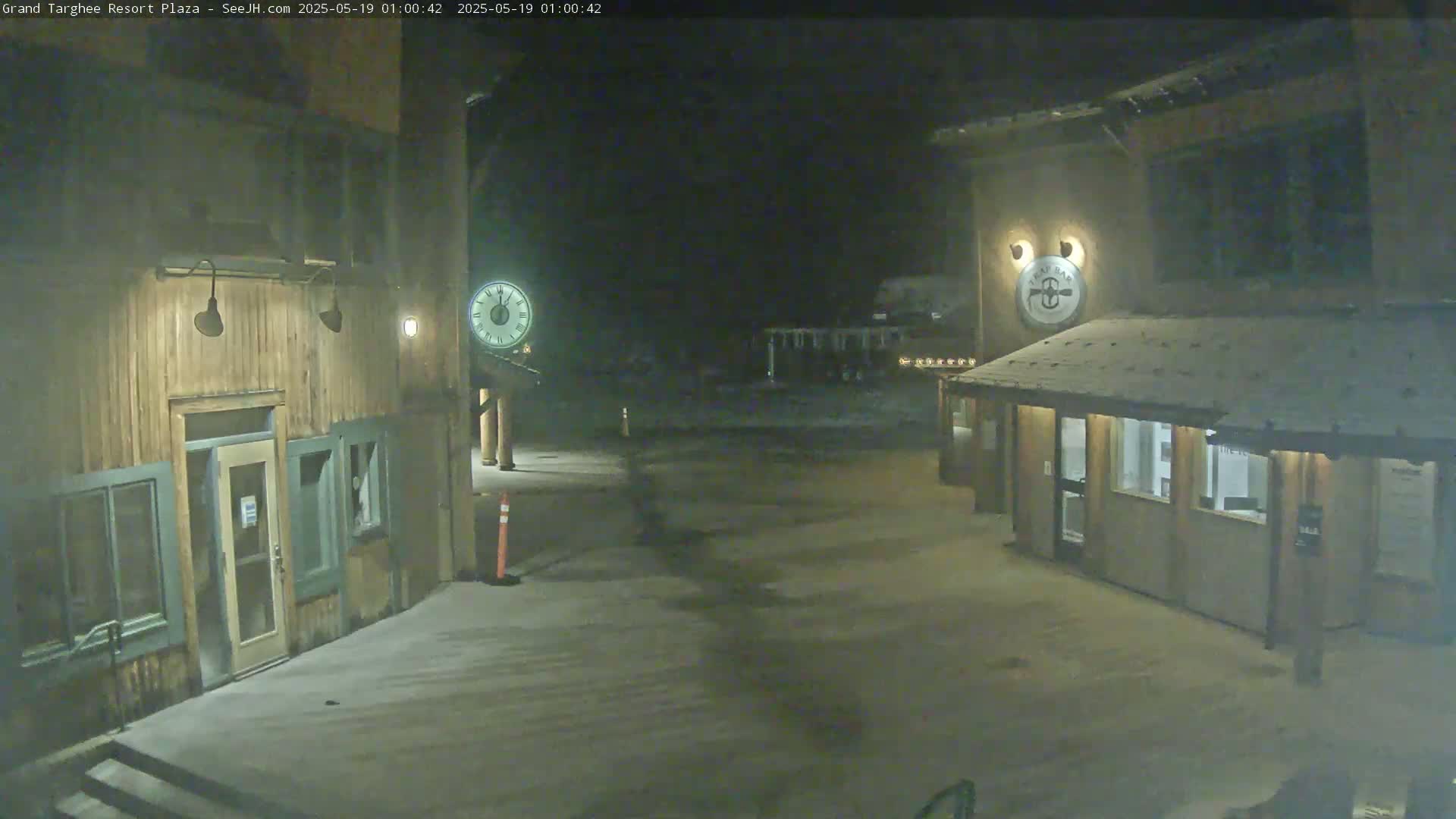 A snow-dusted plaza at night is flanked by two wooden buildings, one with a clock and the other with a circular sign.