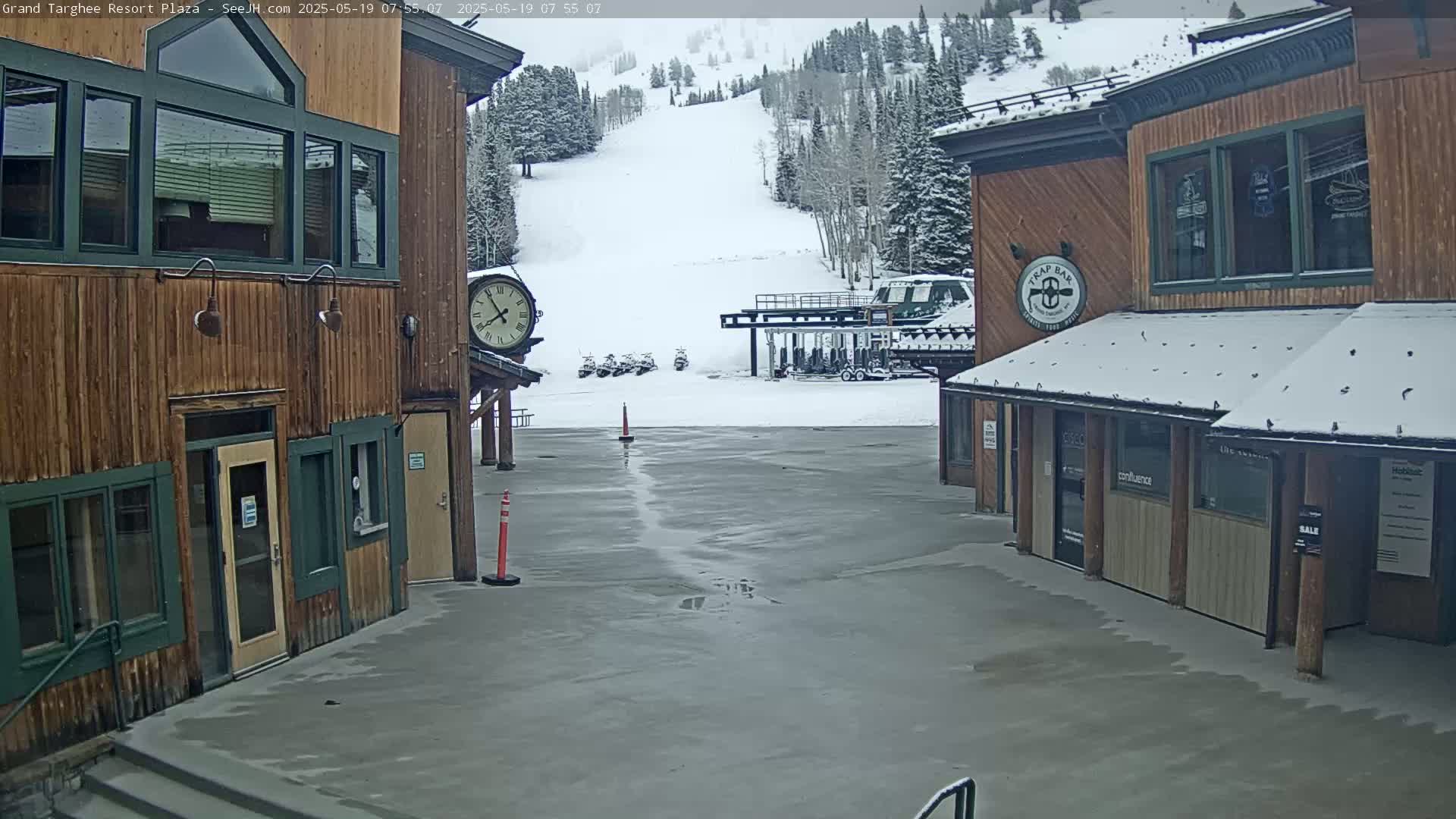A plaza with two wooden buildings, a snow-covered ski slope in the background, and a wet, partially snow-covered ground.