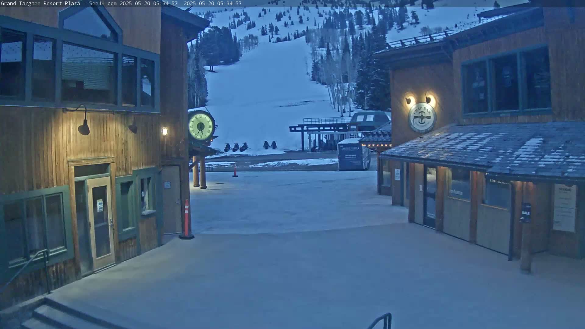 A snow-covered plaza sits between two wooden buildings at twilight, with a ski slope visible in the background.