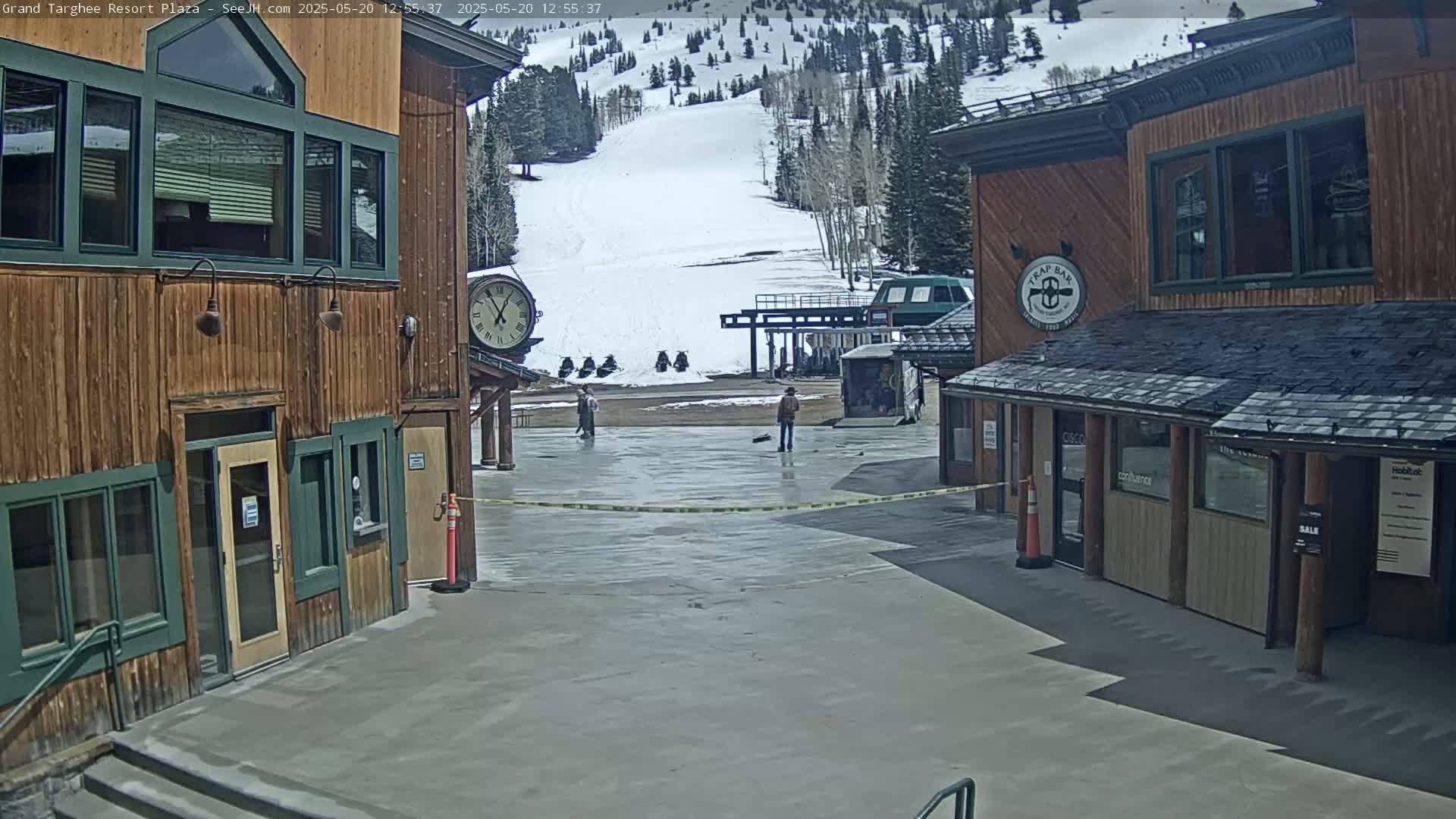 A plaza with two wooden buildings, a large clock, and a snow-covered mountainside in the background under a mostly sunny sky.