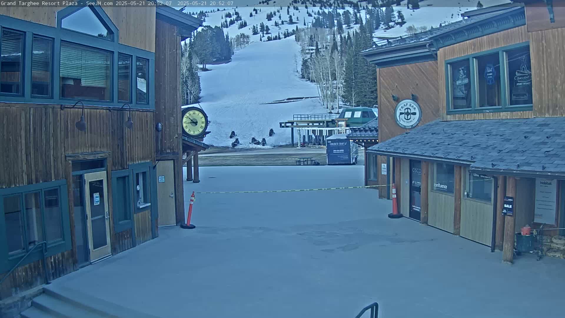 A snow-covered plaza with two wooden buildings sits in front of a ski slope under an overcast sky.