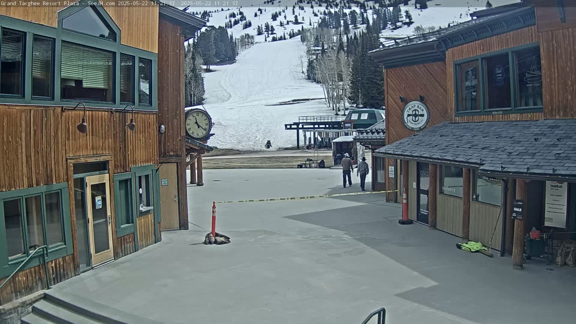 A plaza with two wooden buildings, one featuring a clock, overlooks a snow-covered ski slope on a sunny day.