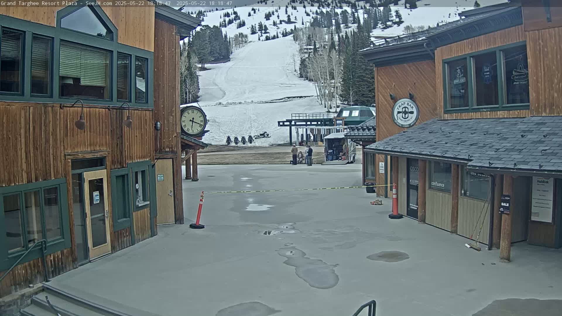 A plaza with two wooden buildings, a snowy mountain in the background, and several puddles on the wet ground.