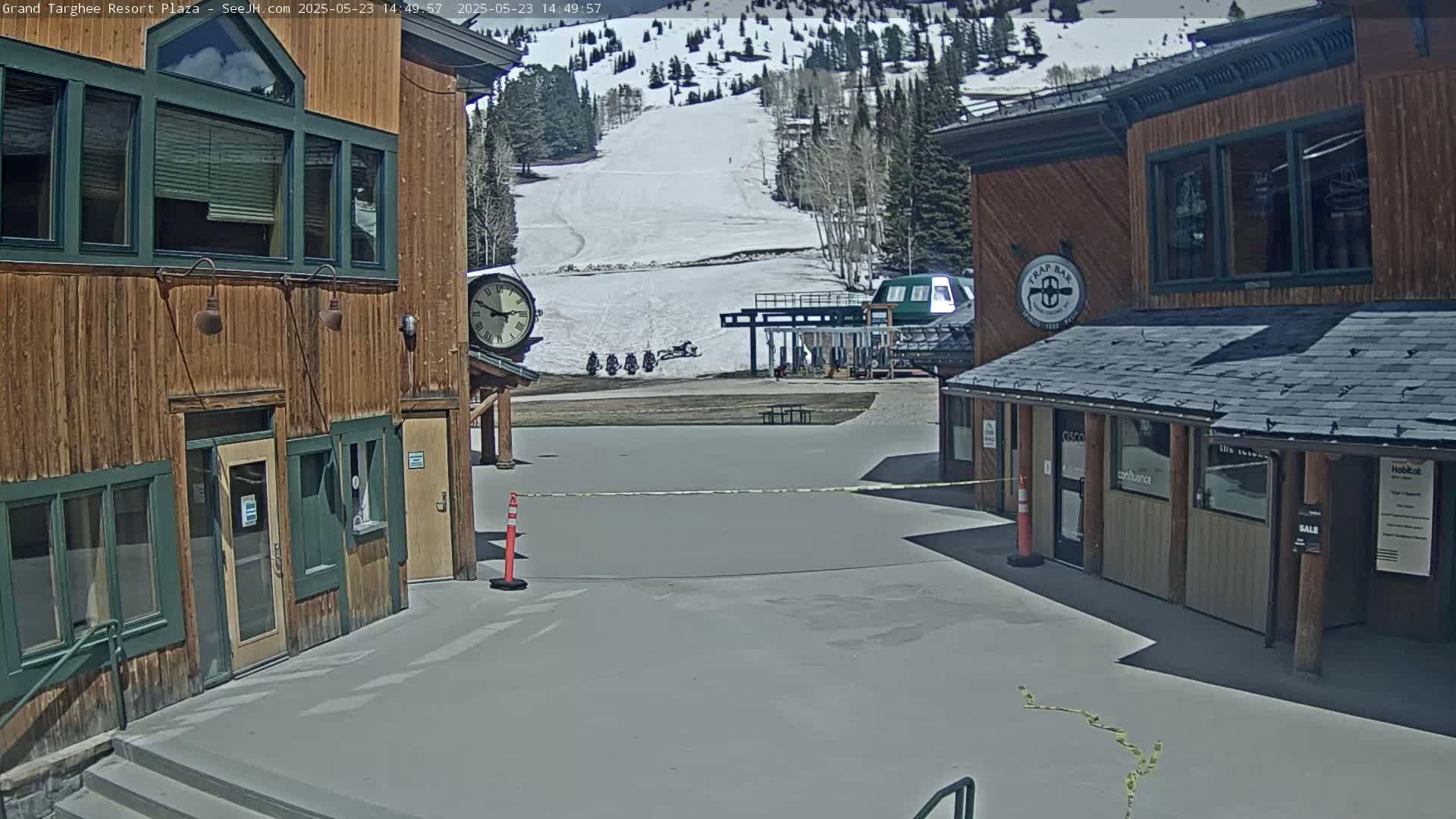 A plaza between two wooden buildings overlooks a snow-covered ski slope on a sunny day, with several snowmobiles parked near a ski lift.